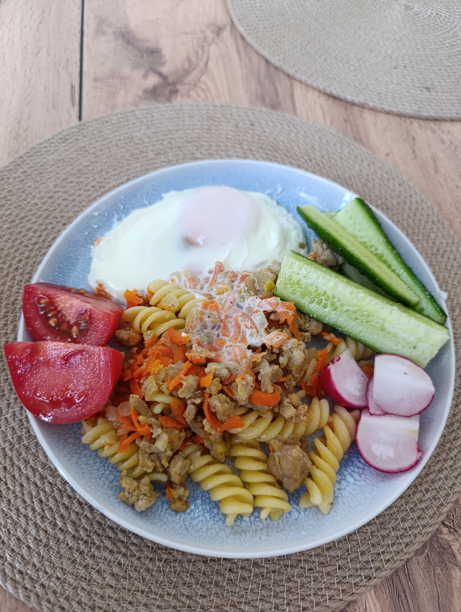 On a light blue plate, a meal consisting of a fried egg, spiral pasta, vegetables (tomato, cucumber, and radish), and a mixture containing ground meat and carrots is presented. The plate rests on a round, brown, woven placemat, which is set on a wooden surface. The scene appears to be indoors.