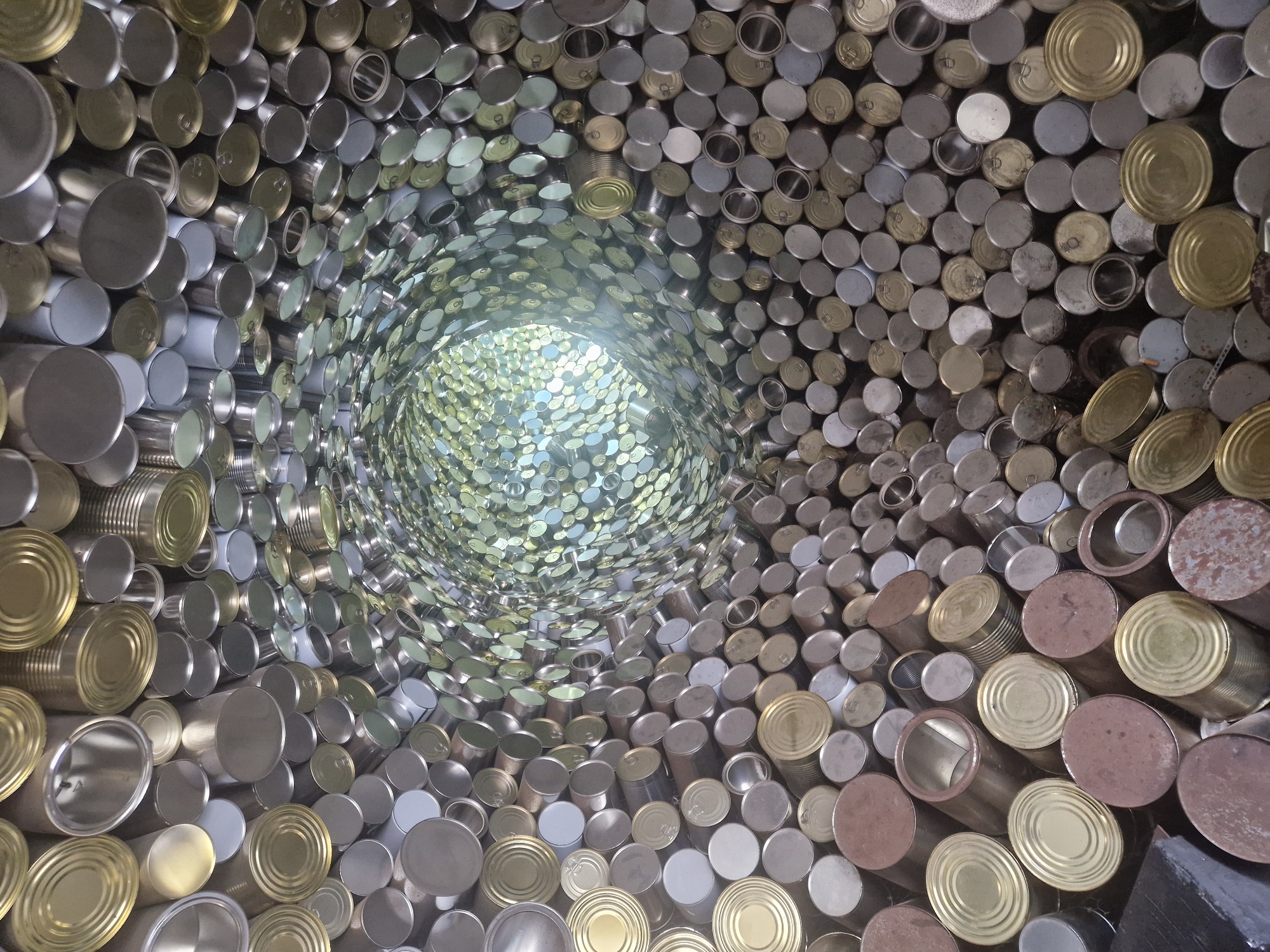 The image depicts an art installation resembling a tunnel constructed entirely of tin cans. The cans vary in color from silver and gold to rusted brown, and are arranged in a circular pattern that recedes into the distance, creating a vortex effect. Light penetrates from the far end of the tunnel. The location is listed as Unknown, Unknown.