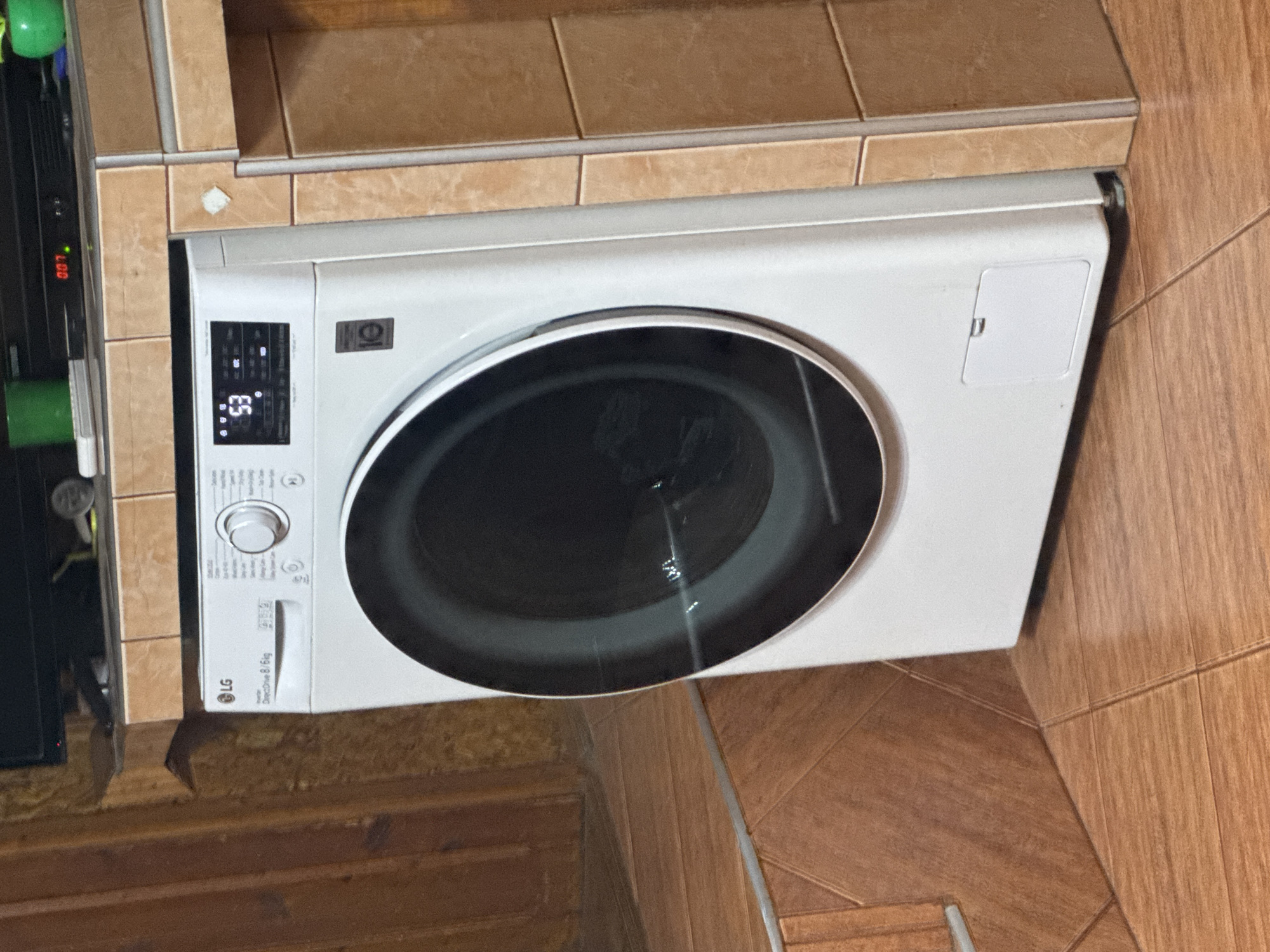 This image, captured in Sarnevo, Bulgaria, displays a domestic interior featuring an LG DirectDrive 8/6kg front-load washing machine positioned in a corner. The washing machine is white, with a prominent dark, circular glass door reflecting ambient light. Its digital control panel displays the number "53" in red LED digits, accompanied by several illuminated white indicator lights. A circular program selection knob is visible on the upper left of the control panel. A "10" logo, indicative of a 10-year warranty, is present on the lower right side of the machine's front panel. A rectangular access panel with a recessed handle is located at the bottom front.
The floor consists of light brown, rectangular ceramic tiles arranged in a staggered pattern with light-colored grout. To the right of the washing machine, a corner structure is covered with smaller, square, light brown ceramic tiles, forming a vertical support and a horizontal countertop. Above the washing machine, on a dark, elevated surface or shelf, a black electronic device is visible, displaying "007" in red LEDs along with a green indicator light. To the left of this device, a green cylindrical object and a white rectangular object are present. To its right, another green object is partially visible. To the left of the washing machine, a wall or structure composed of vertical wooden planks with a light brown finish is observed. No individuals are present in the scene.