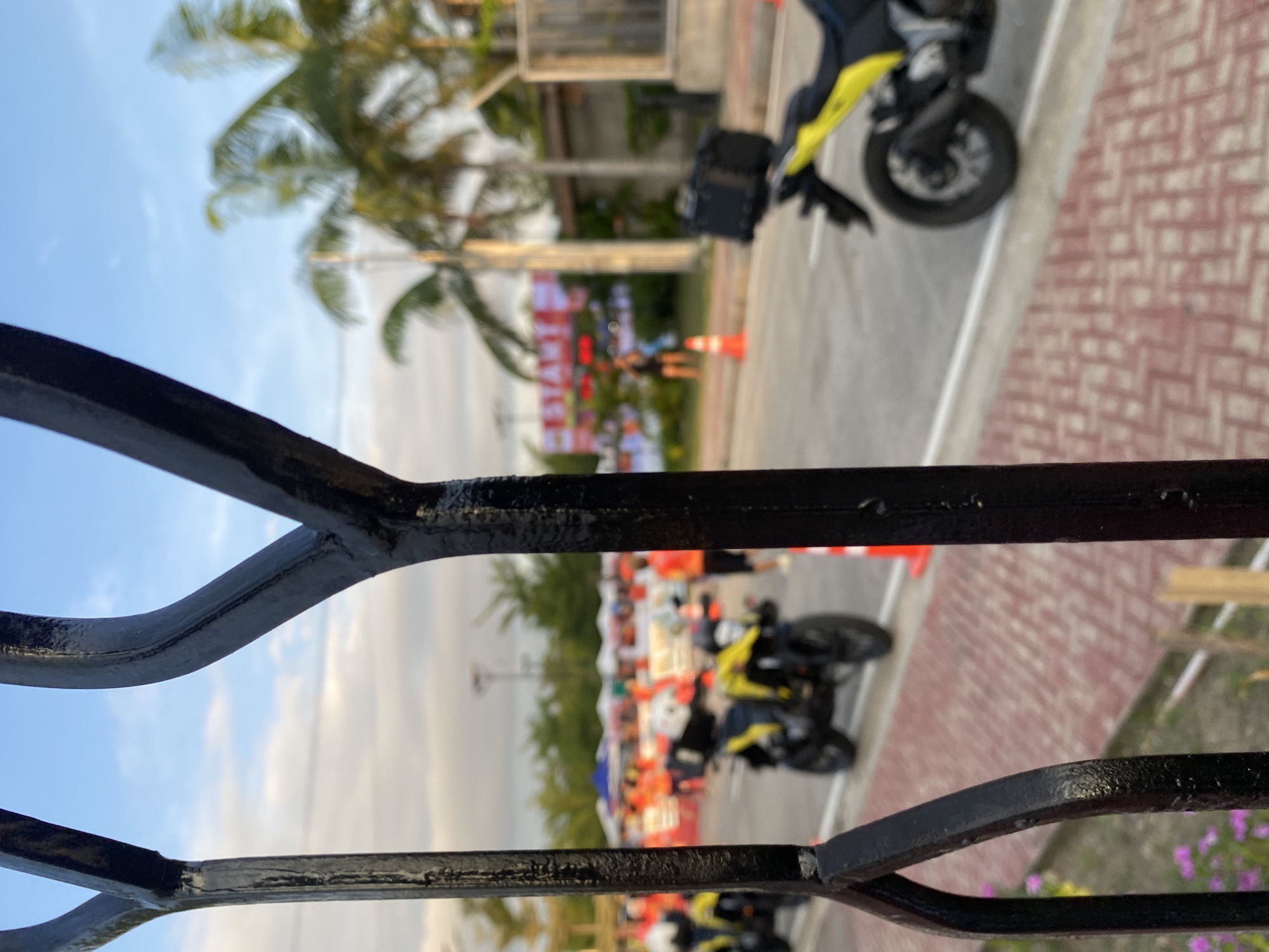 This is a photograph taken from behind a black metal fence, looking out at a street scene in Davao City, Philippines. In the foreground, the black metal bars of the fence are clearly visible, with a patch of pink flowers to the left. Beyond the fence, the scene is slightly blurred due to the focus being on the fence.
The street appears to be set up for an event. There are several yellow motorcycles parked on the road, some with black storage boxes on the back. People in bright orange shirts are visible in the background, suggesting they are participants or staff for an organized event. There are also several white tents set up along the side of the road.
Further in the background, a large red banner with the word "START" in white letters is visible, indicating this is likely a starting line for a race or event. Palm trees are scattered throughout the scene, typical of the tropical climate of Davao City. The sky is partly cloudy, with a mix of blue sky and white clouds. The lighting suggests it is either early morning or late afternoon. The ground appears to be paved with red brick-like material in some areas and asphalt in others. Orange traffic cones are placed along the road.