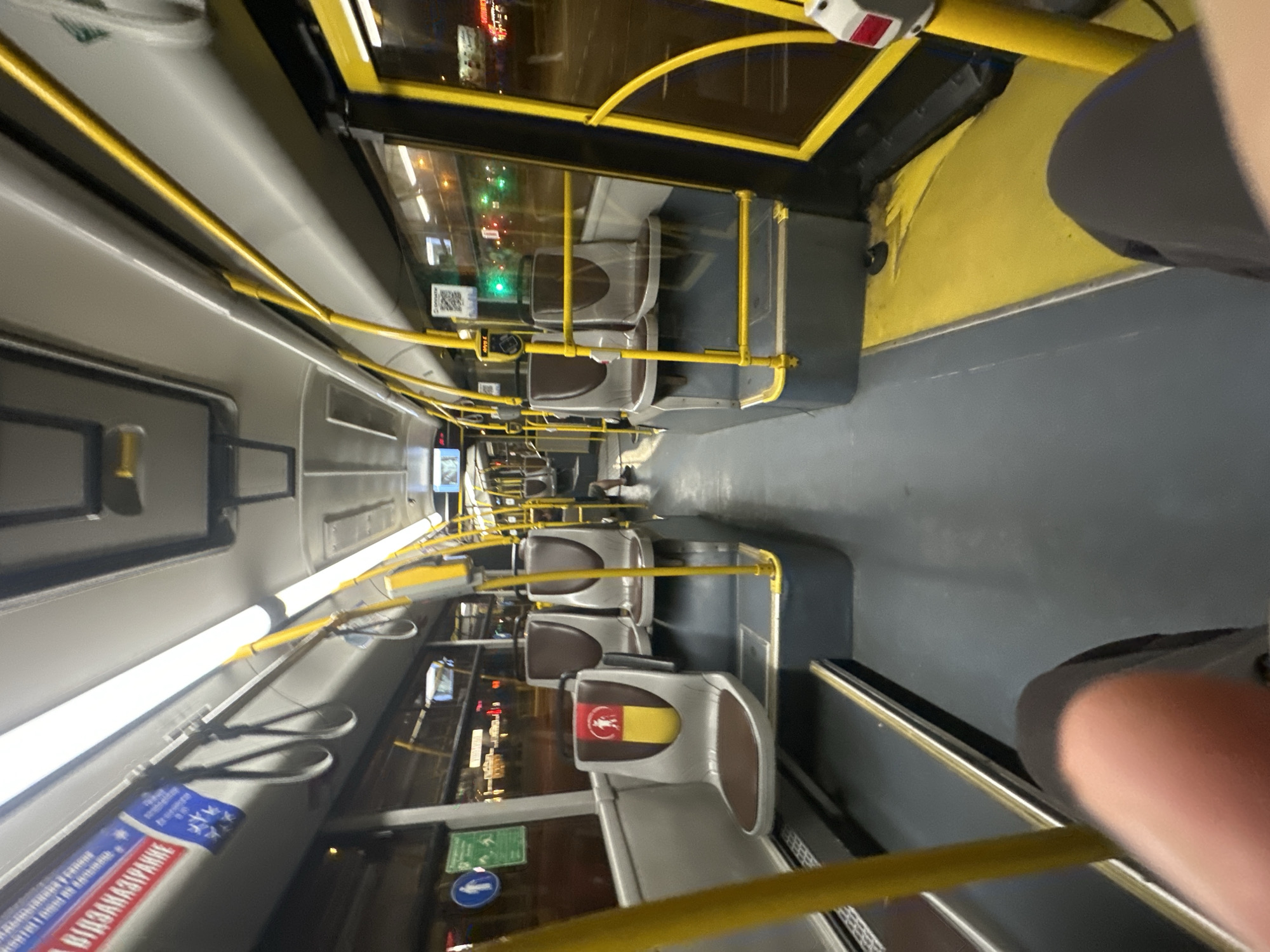 The image shows the interior of a public transport vehicle, likely a bus, in Minsk, Belarus. The perspective is from a seated passenger looking forward. The aisle is visible, with rows of seats on either side. The seats are a neutral gray with brown upholstery. Yellow handrails and poles are prominent throughout the cabin, a common feature in public transportation for passenger support.
On the left side, a seat with a designated priority seating symbol is visible. This symbol features a silhouette of a person with a cane, colored red and yellow. Further forward on the left, there are signs on the ceiling, one of which displays Cyrillic text, possibly indicating bus route information or regulations.
The flooring is a dark gray with yellow accents on the edges and some raised platforms. The windows show a nighttime urban scene with lights and reflections, consistent with a city environment like Minsk. There are screens mounted on the ceiling, typical for displaying route information or advertisements.
In the background, on the right side of the aisle, another passenger is partially visible, seated and wearing dark clothing. The overall lighting inside the bus is bright, likely from fluorescent or LED fixtures on the ceiling. The image captures a mundane moment within the daily commute on public transport in Minsk.