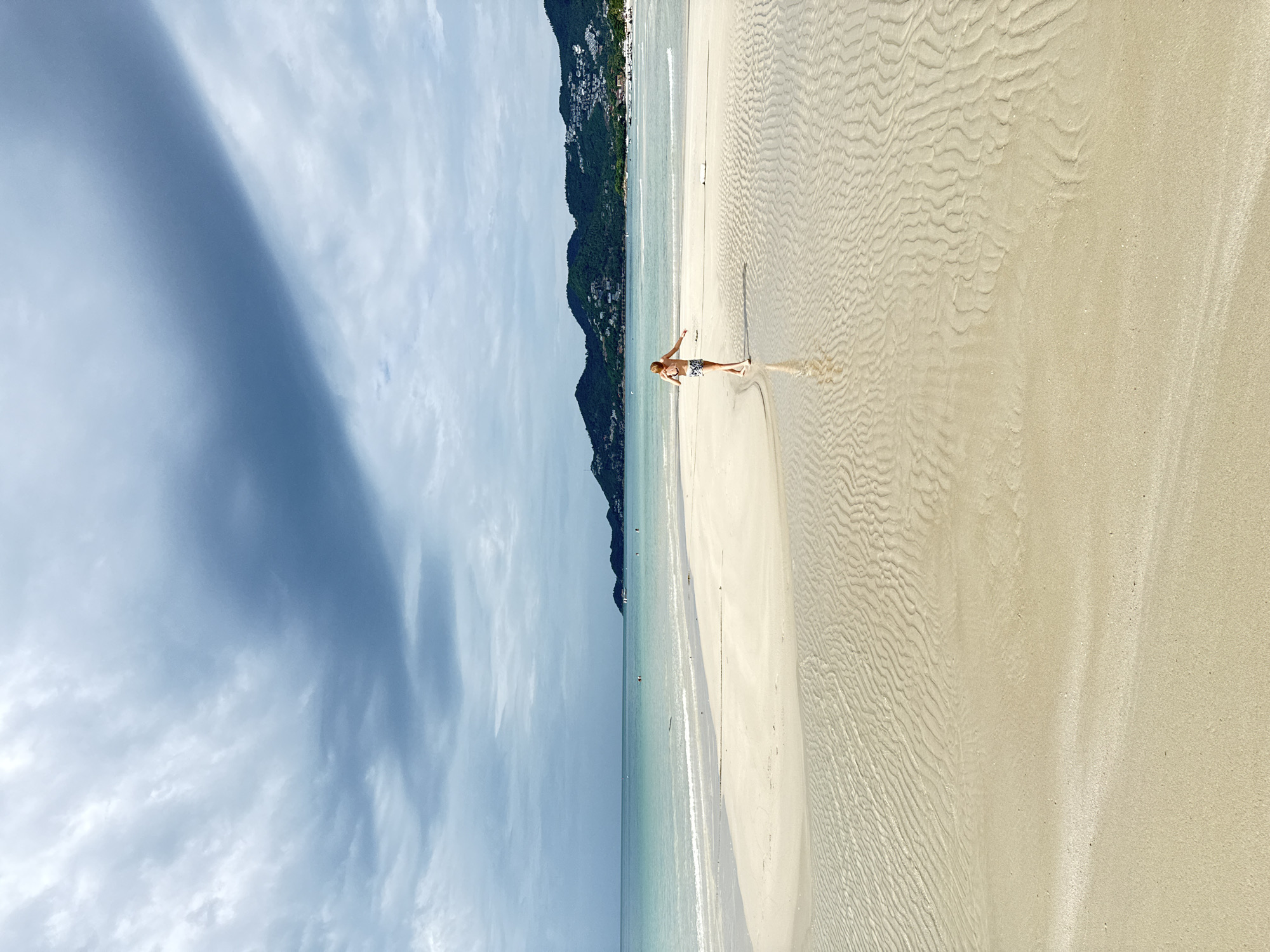 A person walks on a wet sandbar on a beach in the city of Ko Samui District, Thailand. The sky is overcast with dramatic clouds. The water is a turquoise blue and calm, with gentle waves rolling onto the shore. The sand is a light tan color and has ripples from the receding tide. In the background, lush green hills dotted with buildings rise from the coast. The person, facing away from the camera, is wearing a patterned swimsuit and has their arms extended.