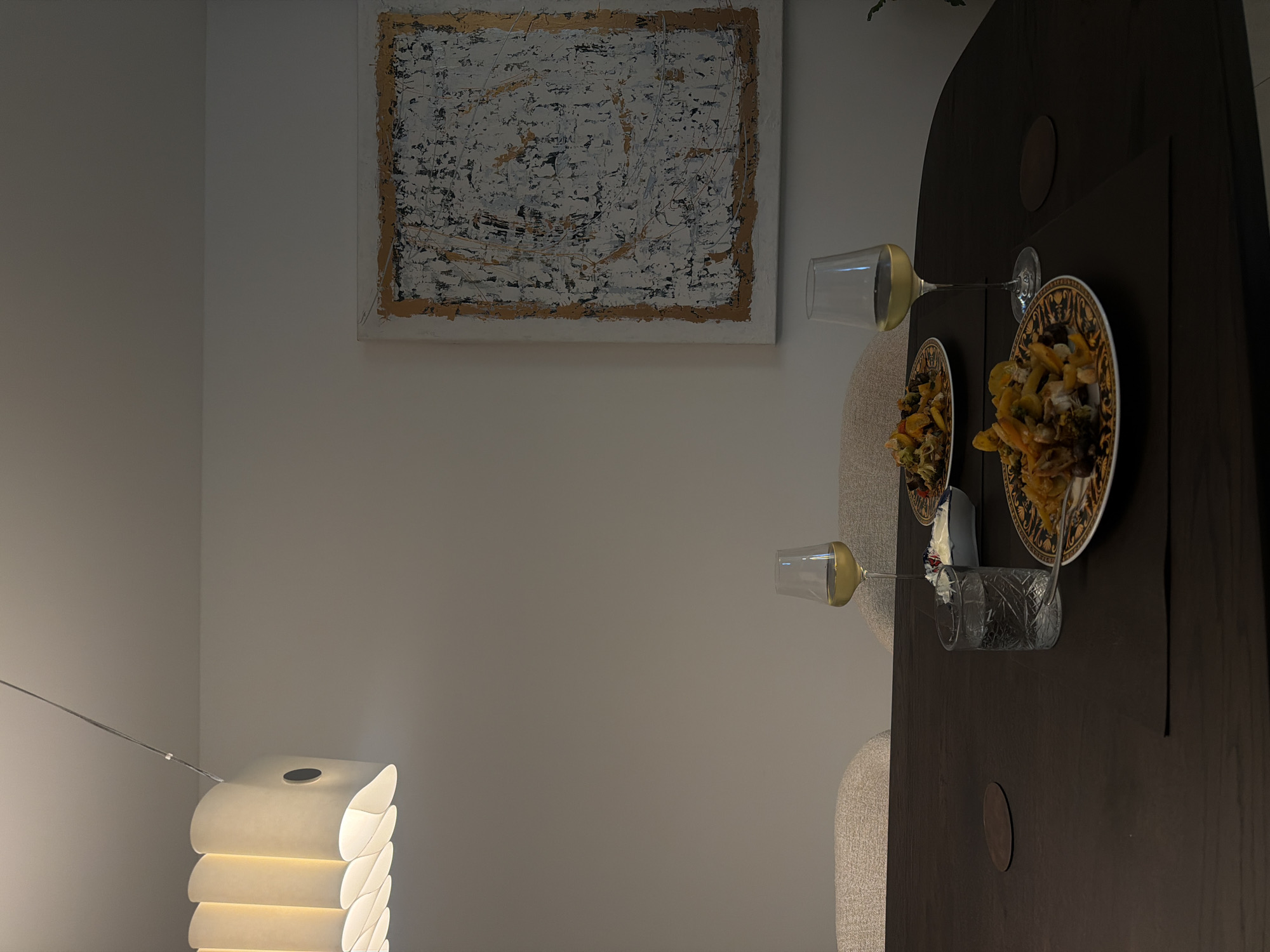 The image depicts a dining scene with two place settings on a dark wood table. Each setting includes a plate of food, a wine glass filled with white wine, and a small dish of what appears to be a creamy dip. A glass of water is also present at one setting. In the background, two chairs with upholstered seats are visible, and a large abstract painting hangs on the wall. A modern pendant light fixture hangs from the ceiling. The lighting in the room is dim, suggesting it might be evening. The scene appears to be set in a dining room or a well-lit area of a home. No people are visible in the image. The image was captured in Ridderkerk, Netherlands.