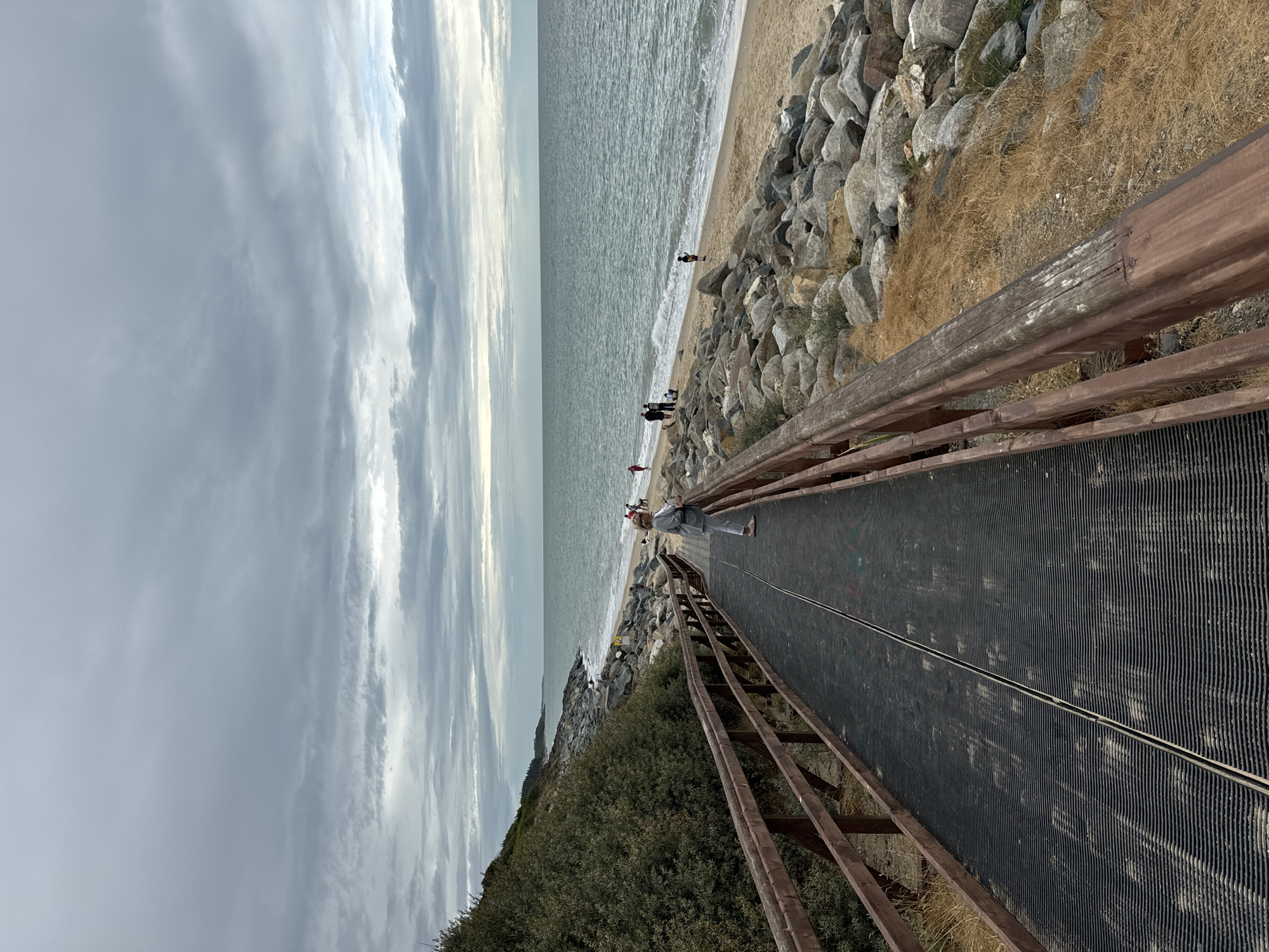 A wooden boardwalk with a black non-slip surface runs diagonally from the bottom left to the middle of the image, leading towards a beach. A woman in a light grey jacket and white pants stands on the boardwalk, facing away from the camera, looking out at the sea. The boardwalk is bordered by wooden railings. To the left of the boardwalk, there is a dense green bush growing on a slight incline. To the right of the boardwalk, there is a sandy beach lined with large grey rocks. The sea, with gentle waves, stretches to the horizon under a cloudy grey sky. Several people are visible on the beach and in the water in the distance. The scene appears to be the boardwalk leading to the beach at Courtown, Ireland.