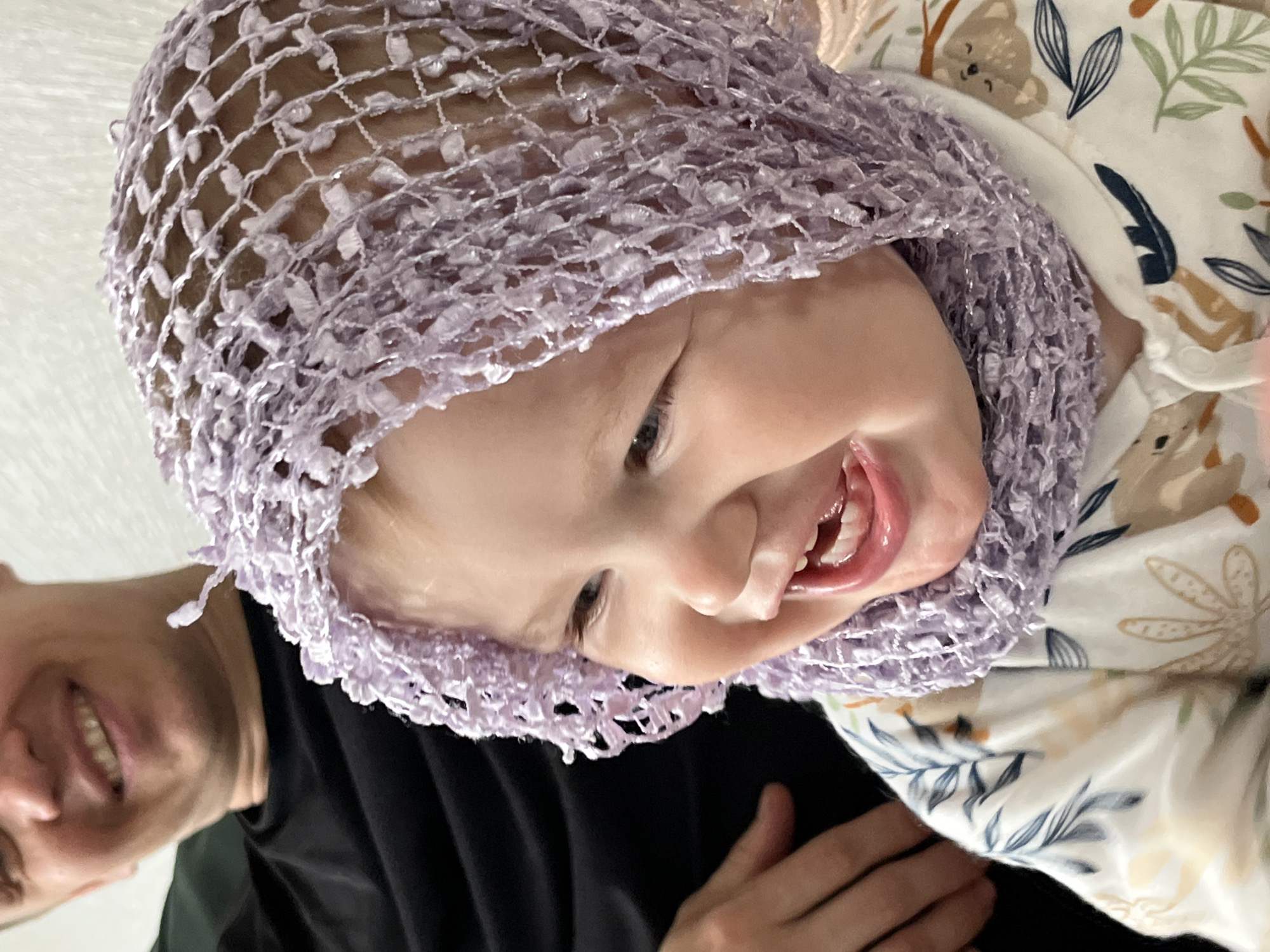The image shows a close-up, tilted view of a baby with a light purple crocheted bonnet. The baby is smiling, revealing some teeth. The baby is wearing a light-colored garment with a print of koalas and leaves. Part of an adult is visible on the left, wearing a black shirt and smiling. The photo was taken in the city of Chaikovsky, Russia.
