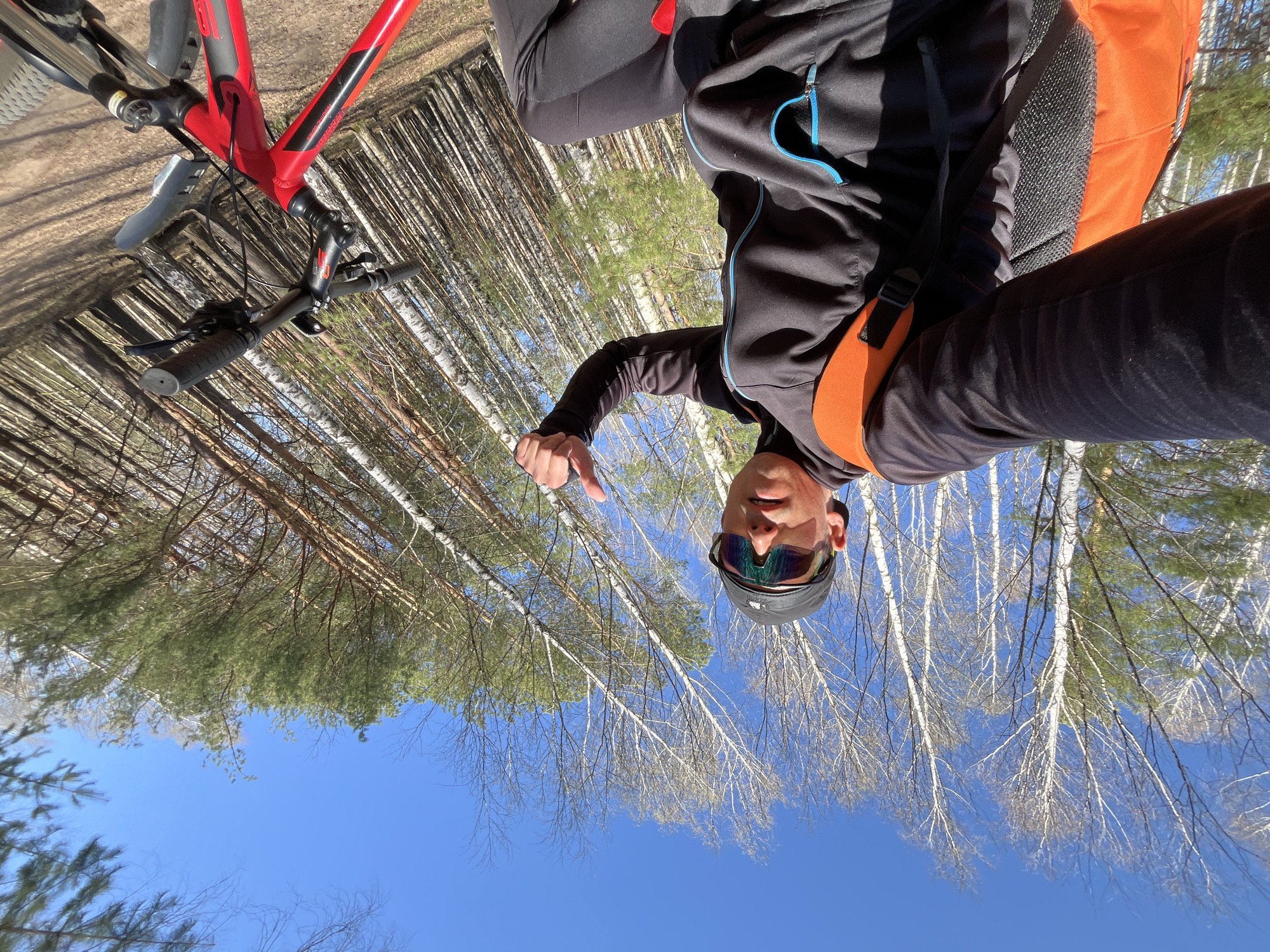 A cyclist stands in a birch forest in Chaikovsky, Russia, next to a red mountain bike and gives a thumbs down. The sky is blue, and the forest has a mix of evergreen and bare birch trees. The cyclist wears dark clothing, sunglasses, and a cap. An orange backpack is visible.