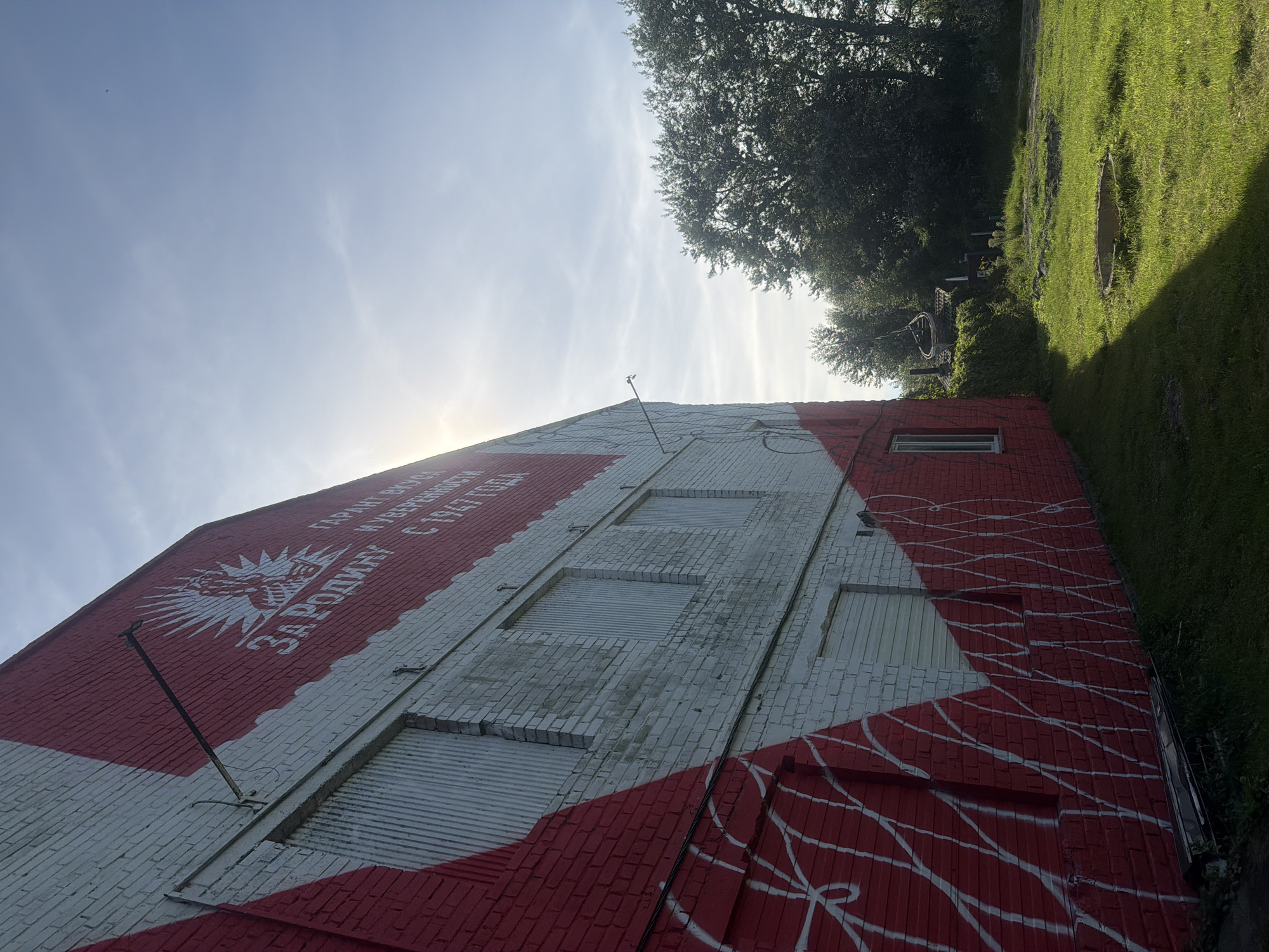 The image captures a brick building in Vzmorye, Russia, adorned with a large mural. The mural is predominantly red and white, with white lettering and a sunburst emblem visible on the upper portion. The building has several windows, some of which appear to be covered with corrugated metal. The exterior walls are made of brick.
The building is situated next to a grassy area with some scattered vegetation and trees in the background. The sky is visible in the upper part of the image, with some clouds and the sun creating a bright glare. There are no people or animals visible in the scene. The overall impression is one of an outdoor, somewhat rural or industrial setting.