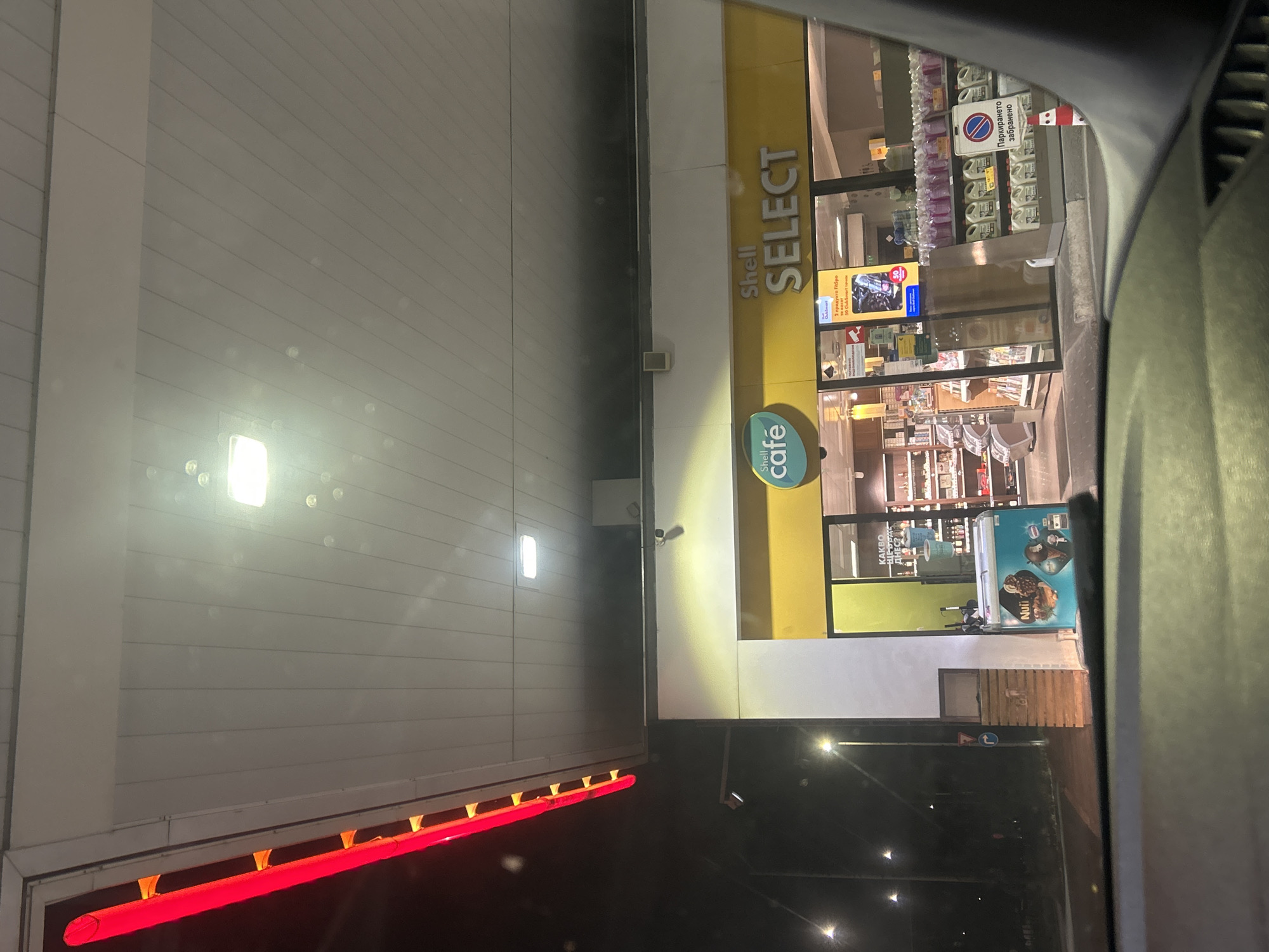 The image shows a Shell Select convenience store at a gas station at night. The store has a yellow awning with the Shell Select logo prominently displayed. Inside, shelves are stocked with various products, and there are display cases for drinks and snacks. A red neon sign runs along the edge of the gas station canopy. Streetlights and interior lights illuminate the scene. The image appears to be taken from inside a vehicle, looking out towards the store. The location is identified as being in Ruse, Bulgaria.