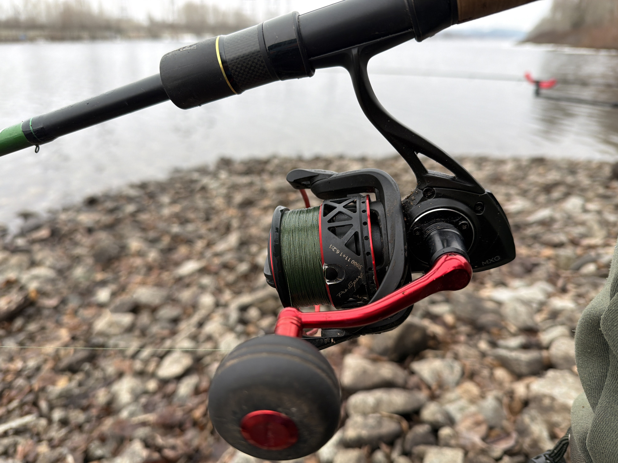 The image features a fishing rod and reel in the foreground, with a rocky shoreline and water in the background. The fishing reel is black with red accents and has green line spooled around it. The fishing rod is black with a cork handle. The shoreline consists of small rocks and pebbles. In the background, a body of water is visible, with a red object floating in the distance. The sky is overcast. The location is the city of Chaikovsky, Russia.