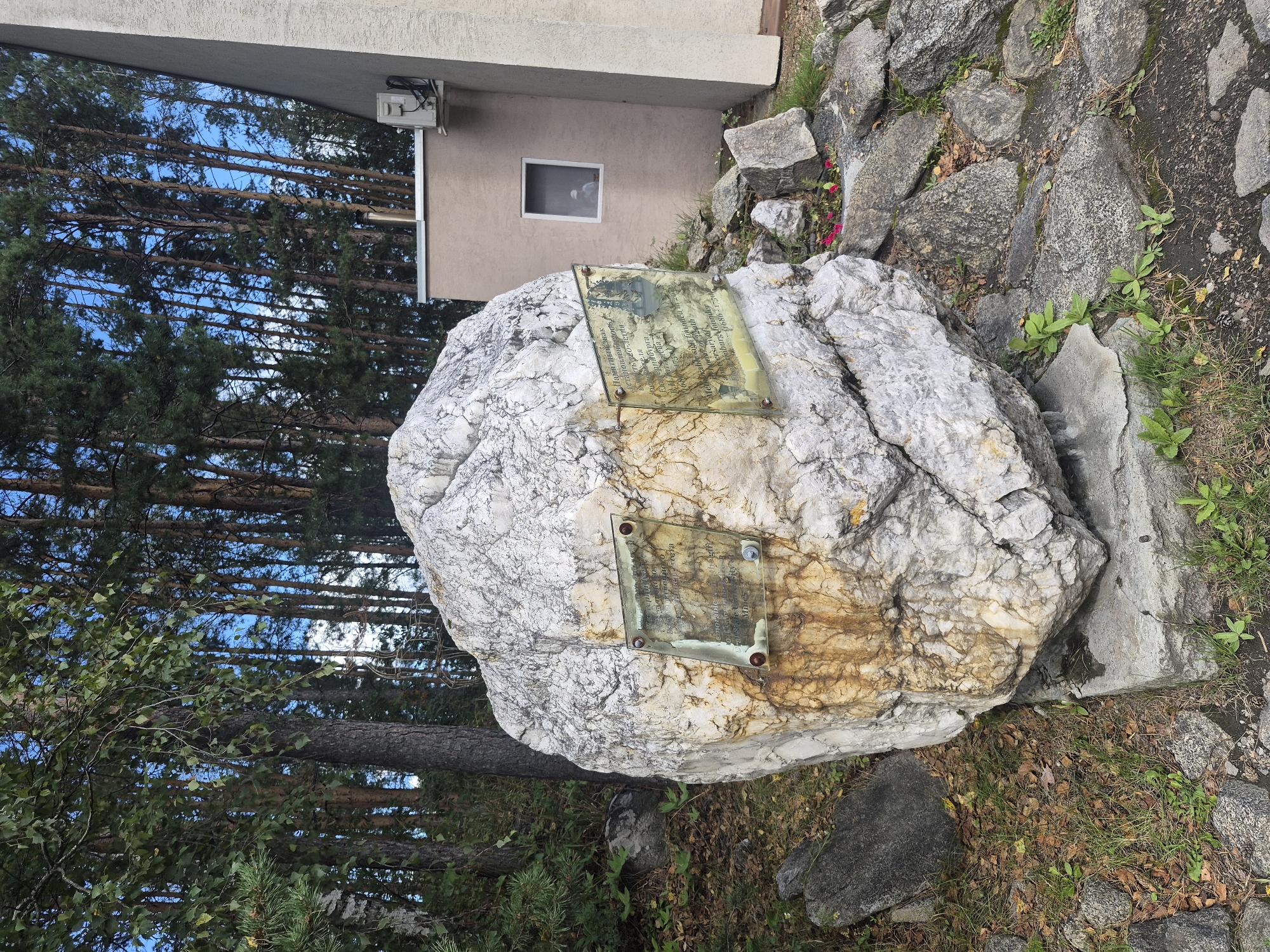 This is a medium shot of a large, rough-textured white boulder with significant rust-colored staining. Two rectangular glass plaques, each with text and an image, are attached to the boulder with metal screws. The boulder is situated outdoors, on a bed of gravel and scattered dry leaves, with smaller rocks and sparse green vegetation around it. In the background, there is a dense forest of tall, slender pine trees with a clear blue sky visible through their branches. To the right of the boulder, a portion of a light-colored building is visible, with a small window and an air conditioning unit mounted on its exterior wall. A small patch of pink flowers is visible near the base of the boulder.
This boulder is located in Yekaterinburg, Russia, near the Church on Blood in honor of All Saints Who Have\
Shone in the Land of Russia, built on the site of the Ipatiev House, where the last Russian emperor Nicholas II and his family were executed. The boulder is a memorial. The text on the plaque is in Russian and English, commemorating the 300th anniversary of the Romanov dynasty and mentioning\
that the founder, Mikhail Romanov, was born in 1596 and died in 1756.