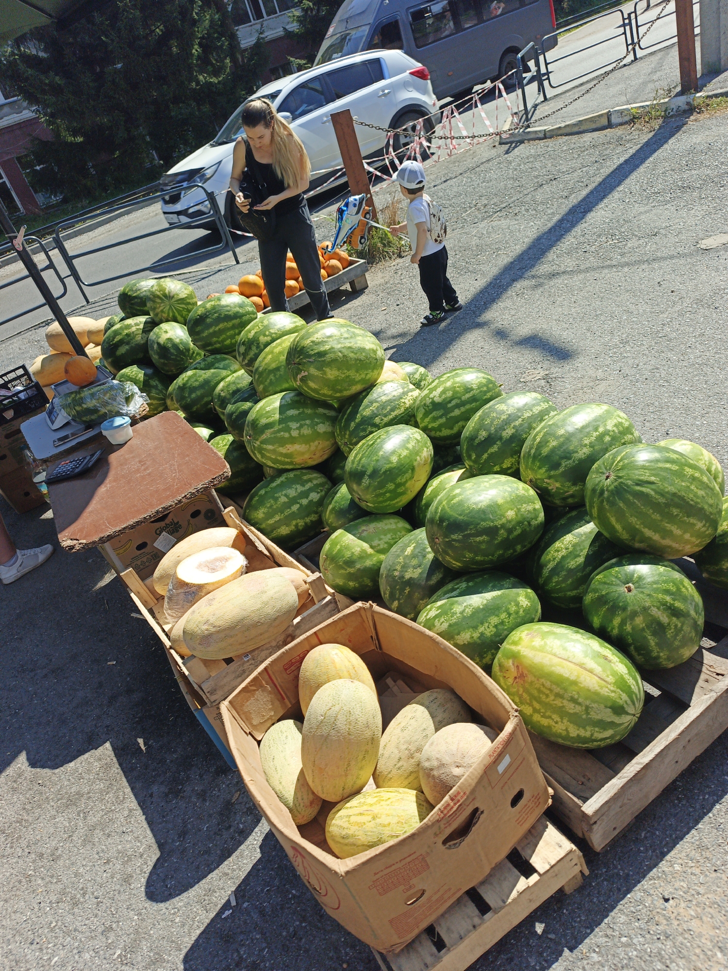 The image depicts an outdoor market scene with a large display of watermelons and melons. A woman with long blonde hair, wearing a black tank top and dark pants, stands behind a table covered with fruits. She is looking down at something in her hand. A young boy in a white t-shirt, dark pants, and a white cap stands nearby, facing away from the camera. In the background, a white SUV and a dark gray van are parked. The market stall is set up on an asphalt surface, with wooden pallets used as stands for the produce. The scene appears to be a street-side vendor stall, possibly in Zlatoust, Russia, given the context provided, under bright sunlight casting long shadows.