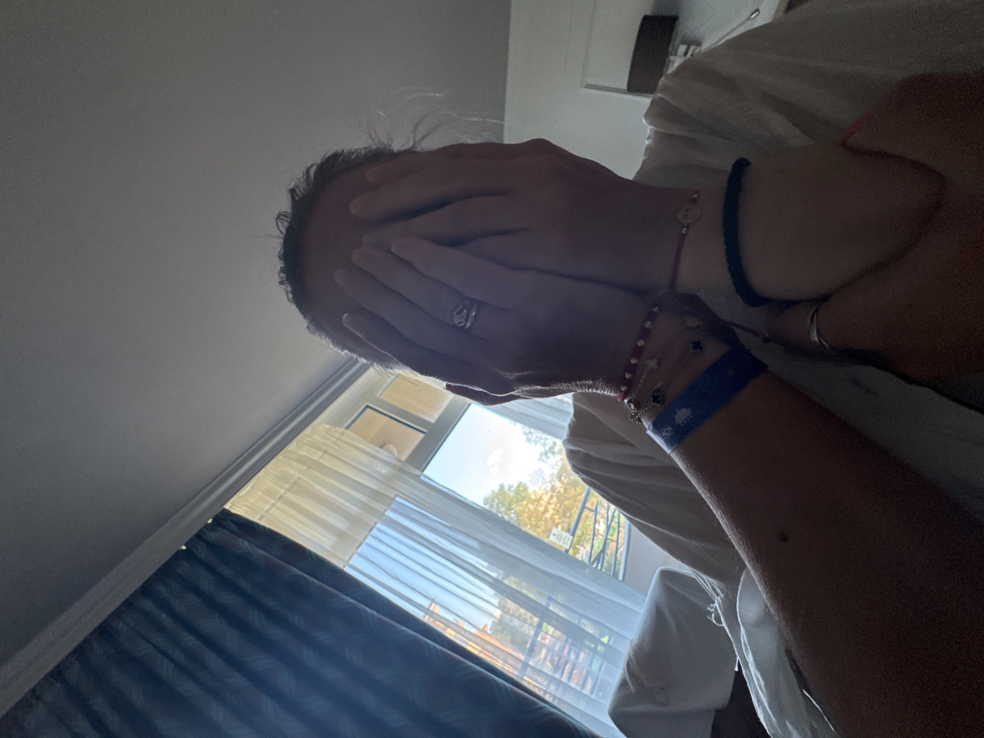 A person is covering their face with their hands while sitting in what appears to be a room. They are wearing several bracelets on their arms, including a blue wristband that says "BEACH." There are dark blue curtains on the left side of the image and sheer white curtains covering a window on the right. Through the window, trees and buildings can be seen, suggesting an outdoor environment. The overall lighting is dim, with light coming from the window. The location is not discernible from the image itself; however, if this scene were in Nesebar, Bulgaria, it could be a hotel room or residential interior overlooking a street or courtyard with typical Bulgarian architecture and vegetation.