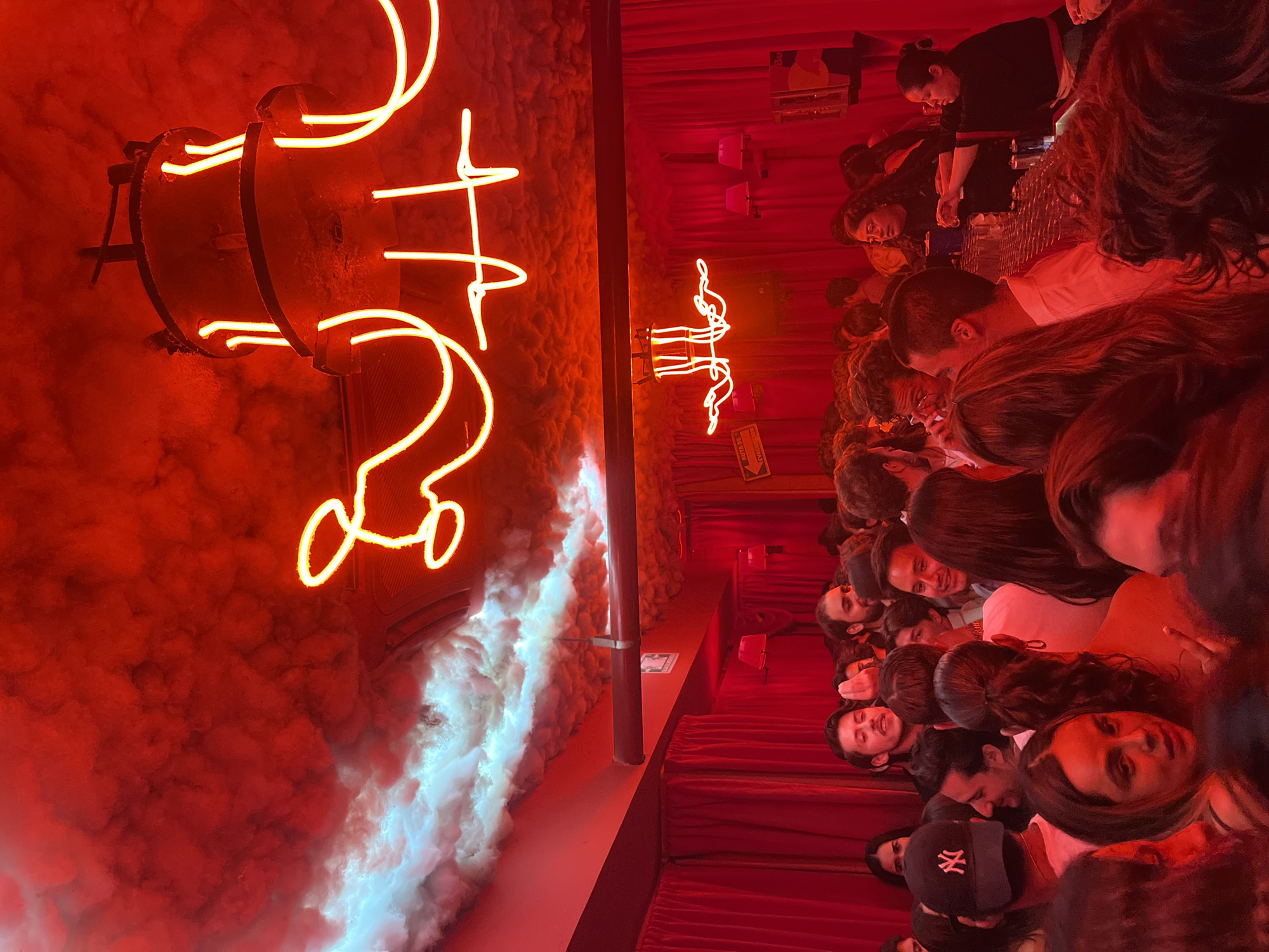 The image shows an indoor view of a crowded bar or club in San Bernardino Tlaxcalancingo, Mexico. The lighting is predominantly red. On the left, neon signs are mounted on a textured wall, with fog effects billowing around them. The right side shows a close-up of a dense crowd, revealing faces of various individuals. A woman in a black uniform is behind a bar, appearing to be working. Rows of seating are visible along the bottom of the image.