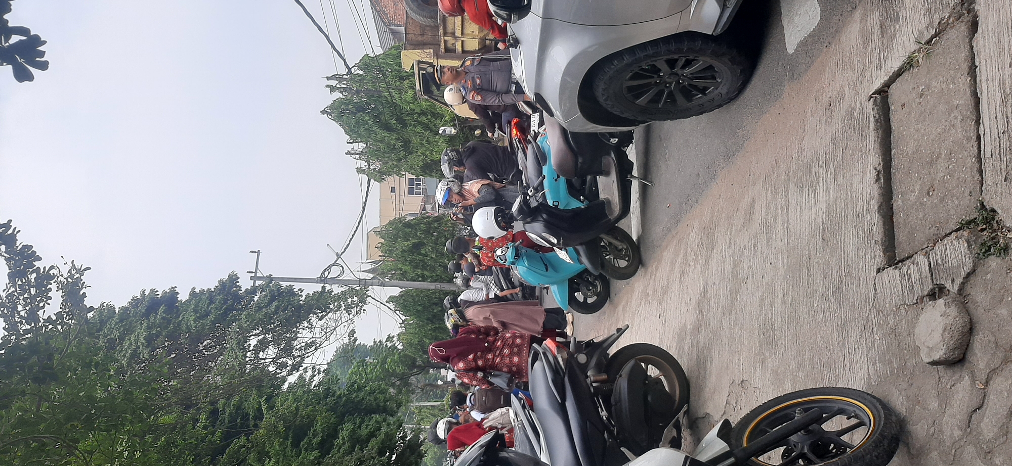 The image captures a street scene in Jambi, Indonesia, likely during daytime with overcast skies. The foreground features the side of a silver SUV, with its front passenger-side wheel and fender visible. To the left, parts of several motorcycles are visible, including a black scooter with a gold rim on its front wheel. The midground is populated by a collection of people and motorcycles. Several individuals, some wearing hijabs, are seen standing or on their scooters. A light blue scooter and a dark scooter are prominently displayed. A man in a grey uniform with orange accents and a cap, possibly a traffic officer, stands near the SUV. Further in the background, more people and a yellow truck are visible. Trees line the left side of the street, and utility poles with wires stretch across the scene. The road surface is paved and shows some wear and tear. The overall impression is of a moderately busy urban street.