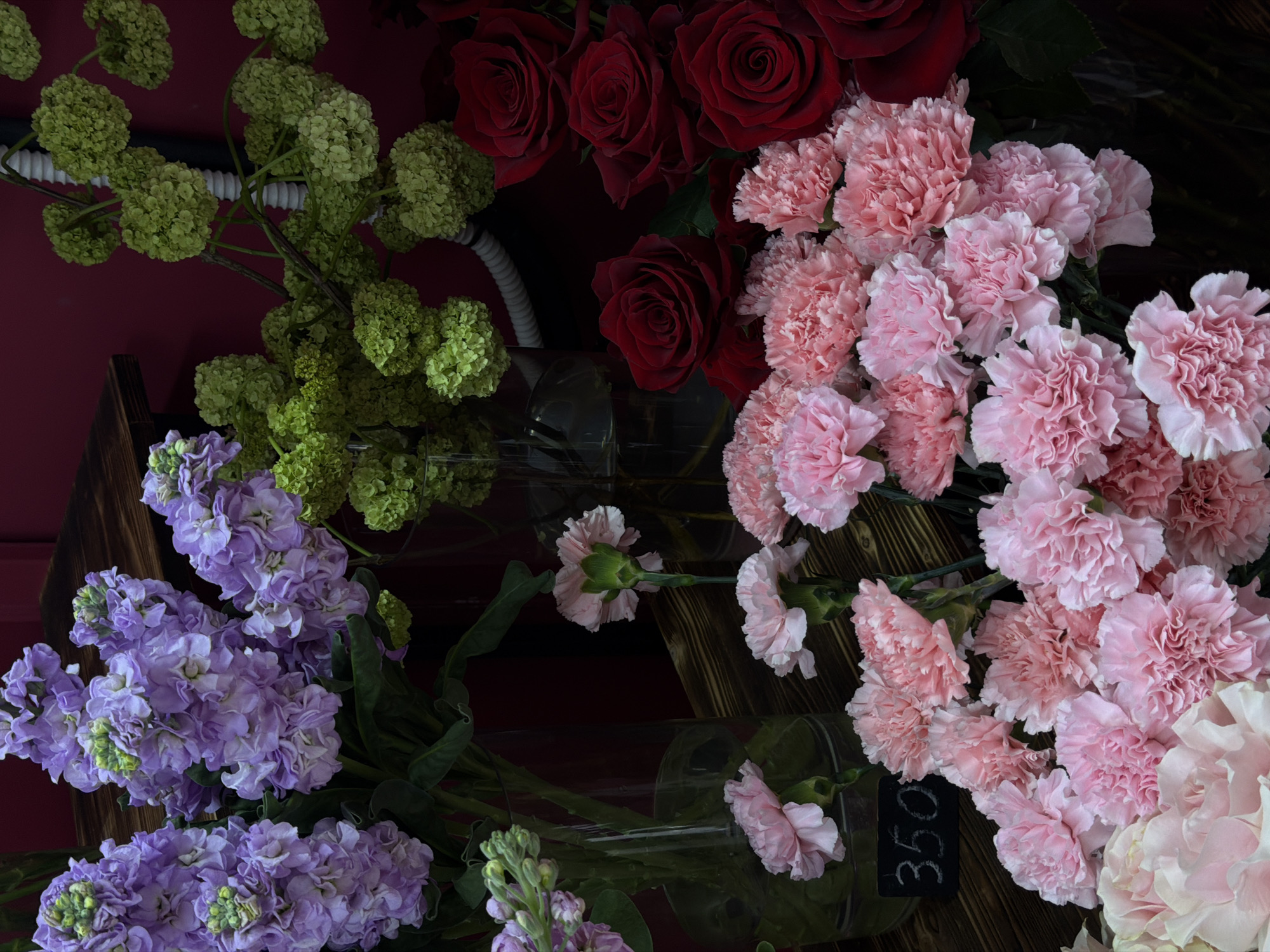 The image shows an assortment of flowers on display, likely for sale, in Krasnoyarsk, Russia. The flowers include red roses, pink carnations, purple stock flowers, and green viburnum. The flowers are arranged in bouquets and possibly displayed in vases on a dark wooden shelf. A small price tag marked "350" is visible near the carnations. The lighting is somewhat dim, suggesting an indoor setting.