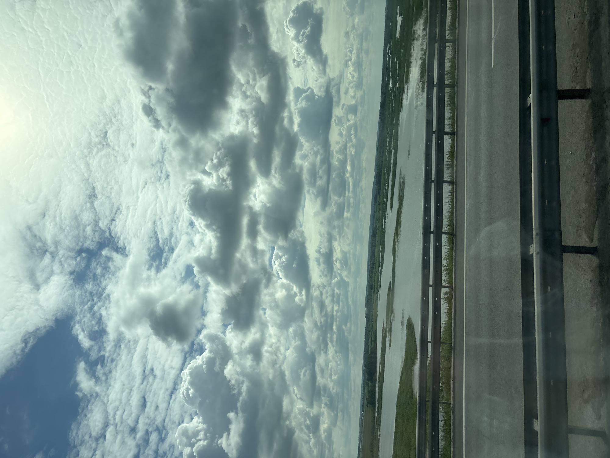 The image shows a wide, calm body of water with green reeds growing along its edges, likely part of the Kama River near Menzelinsk, Russia. In the foreground, a highway runs parallel to the water, separated by metal guardrails. The sky is filled with dramatic, volumetric clouds, some of which are dark and shadowed, suggesting a dynamic weather system. The sun appears to be shining through breaks in the clouds, casting a bright, diffused light. There are no people or vehicles visible in the image, giving the scene a sense of natural beauty and tranquility. The overall impression is of a vast, open landscape under a cloudy sky.