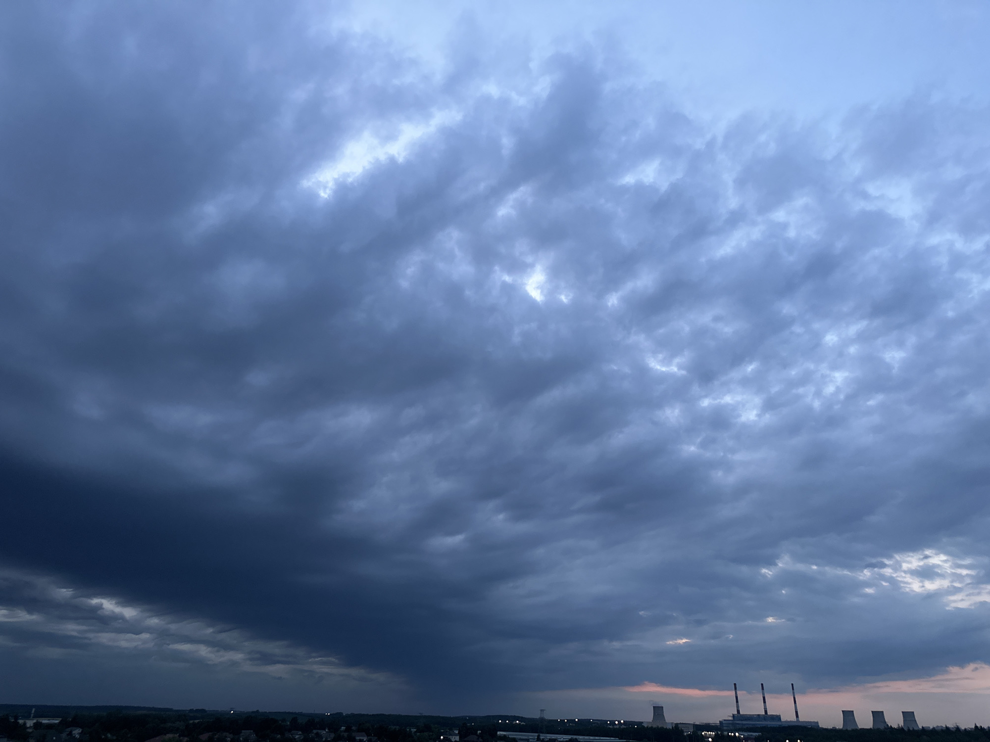 The image depicts a dramatic sky filled with dark, heavy clouds over a landscape that includes a city or town. In the distance, several industrial chimneys, likely part of a power plant or factory, are visible. The sky transitions from a deep, ominous blue-grey on the left, suggesting an approaching storm, to lighter, more scattered clouds towards the right. The lower portion of the image shows a dark, silhouetted horizon with faint lights from buildings and the industrial structures. The overall impression is one of impending weather changes. The scene is located in the vicinity of Шчомысліцкі сельскі Савет, Belarus.