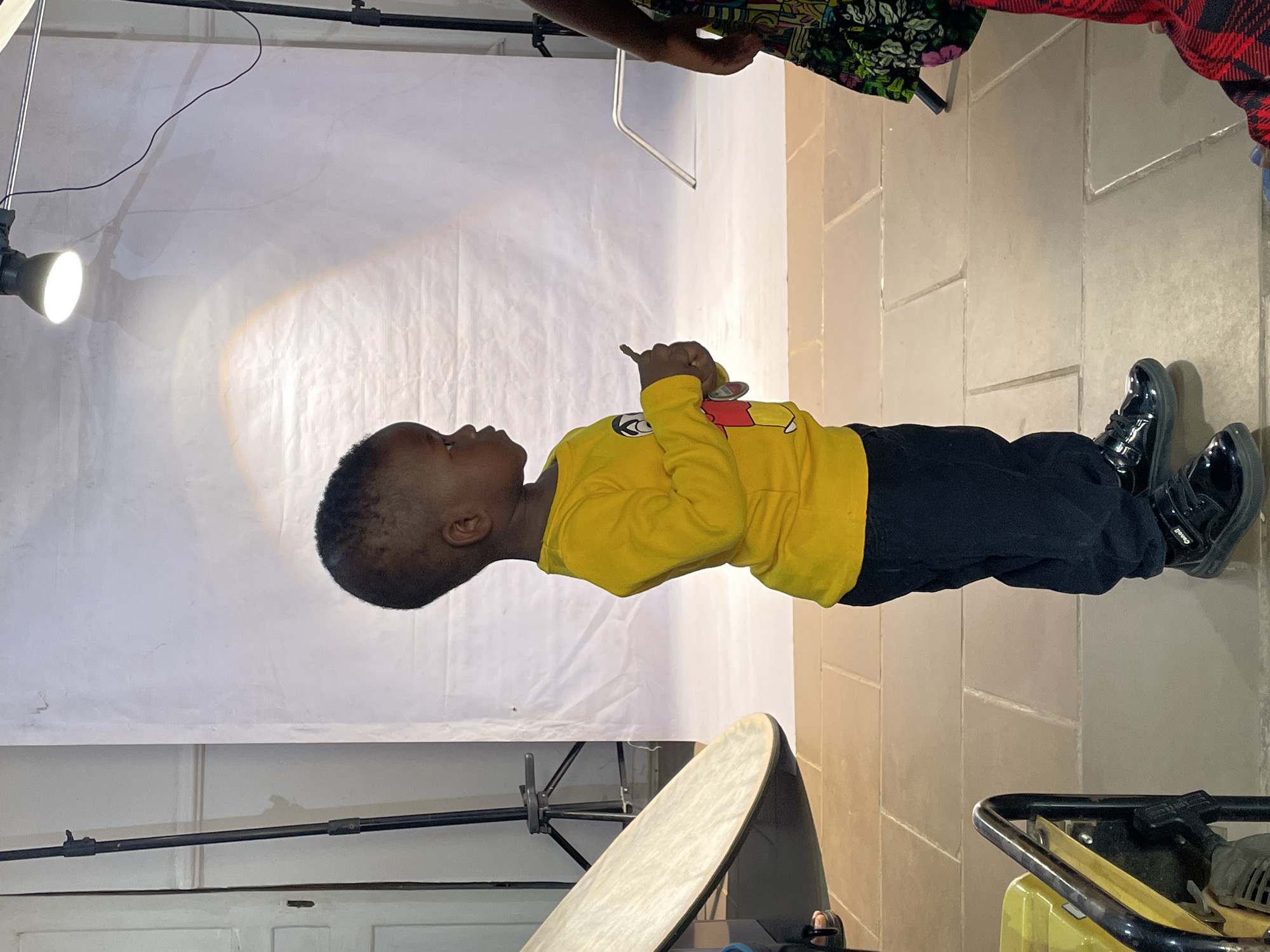 A young Black child stands facing left in an indoor setting, likely a photography studio. The child wears a bright yellow long-sleeved shirt with a red and white cartoon emblem on the chest, black trousers, and shiny black patent leather-like shoes. The word "Castell" is visible on the strap of one shoe. The child holds a small, thin stick-like object in their right hand and a metallic ring with a small charm or key in their left hand, looking slightly upwards with a neutral expression.
The background consists of a plain white, somewhat wrinkled fabric backdrop. The floor is covered with large, light-colored square tiles. To the upper right, an adult's hand and arm, dressed in a colorful patterned fabric, are partially visible, alongside a glimpse of red and black plaid fabric.
Studio equipment is present, including a bright spotlight on a stand on the left, a silver photography reflector disc propped on the lower left, and a partially visible yellow and black piece of machinery (possibly a generator) on the lower right. Black wires are seen stretching across the upper part of the scene. The overall scene suggests a photoshoot or a posed moment for the child in a prepared environment. The location is Jos, Nigeria.