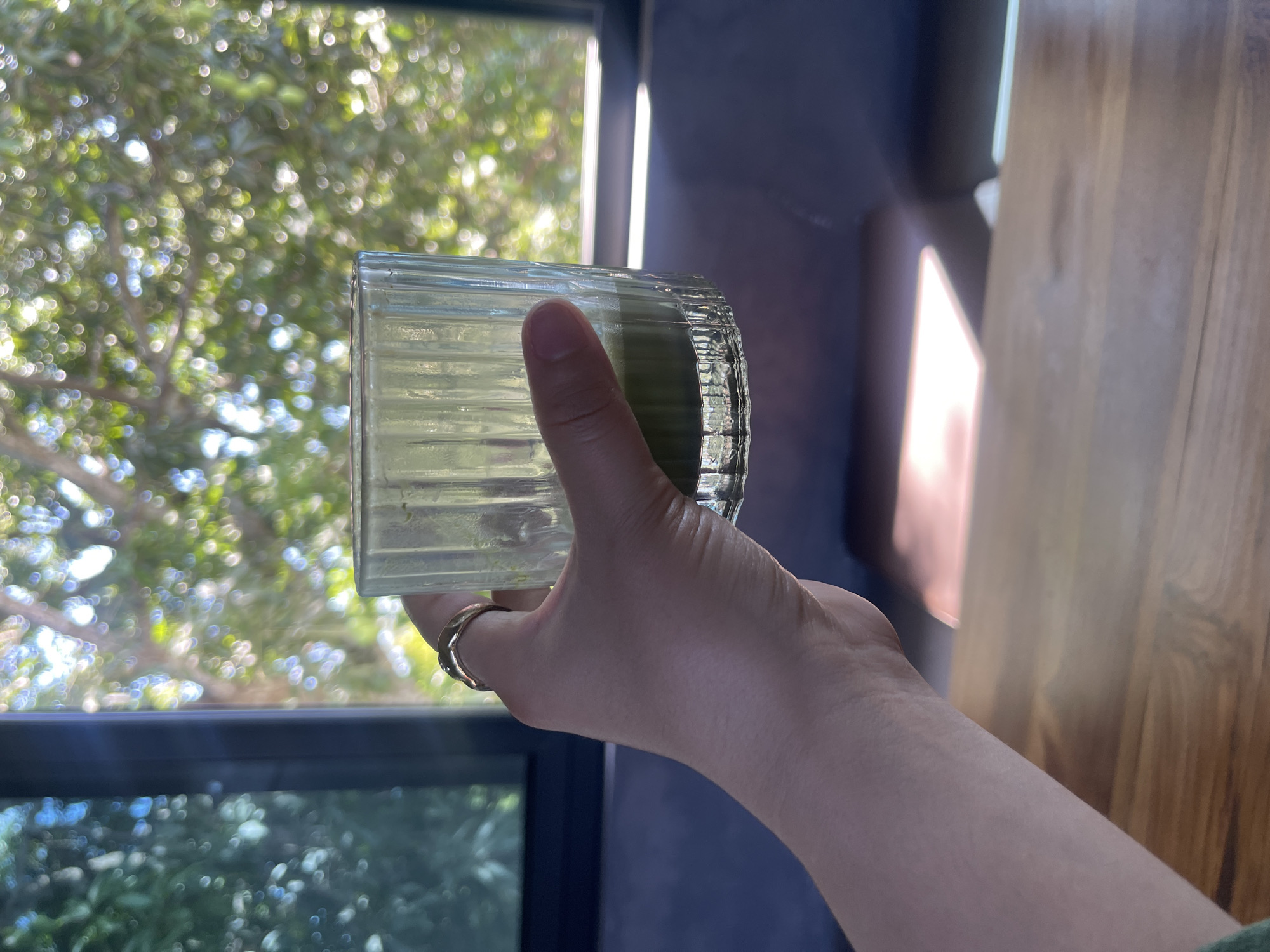A hand wearing a gold ring is holding a glass of green liquid. The glass has vertical ridges and is about one-third full. The hand is positioned in front of a window with a blurred view of green foliage and sunlight filtering through. The scene appears to be indoors, possibly in a cafe or restaurant, with a wooden table in the foreground. The background suggests an outdoor environment with lush vegetation, indicating a tropical location like Kuta Selatan, Indonesia.