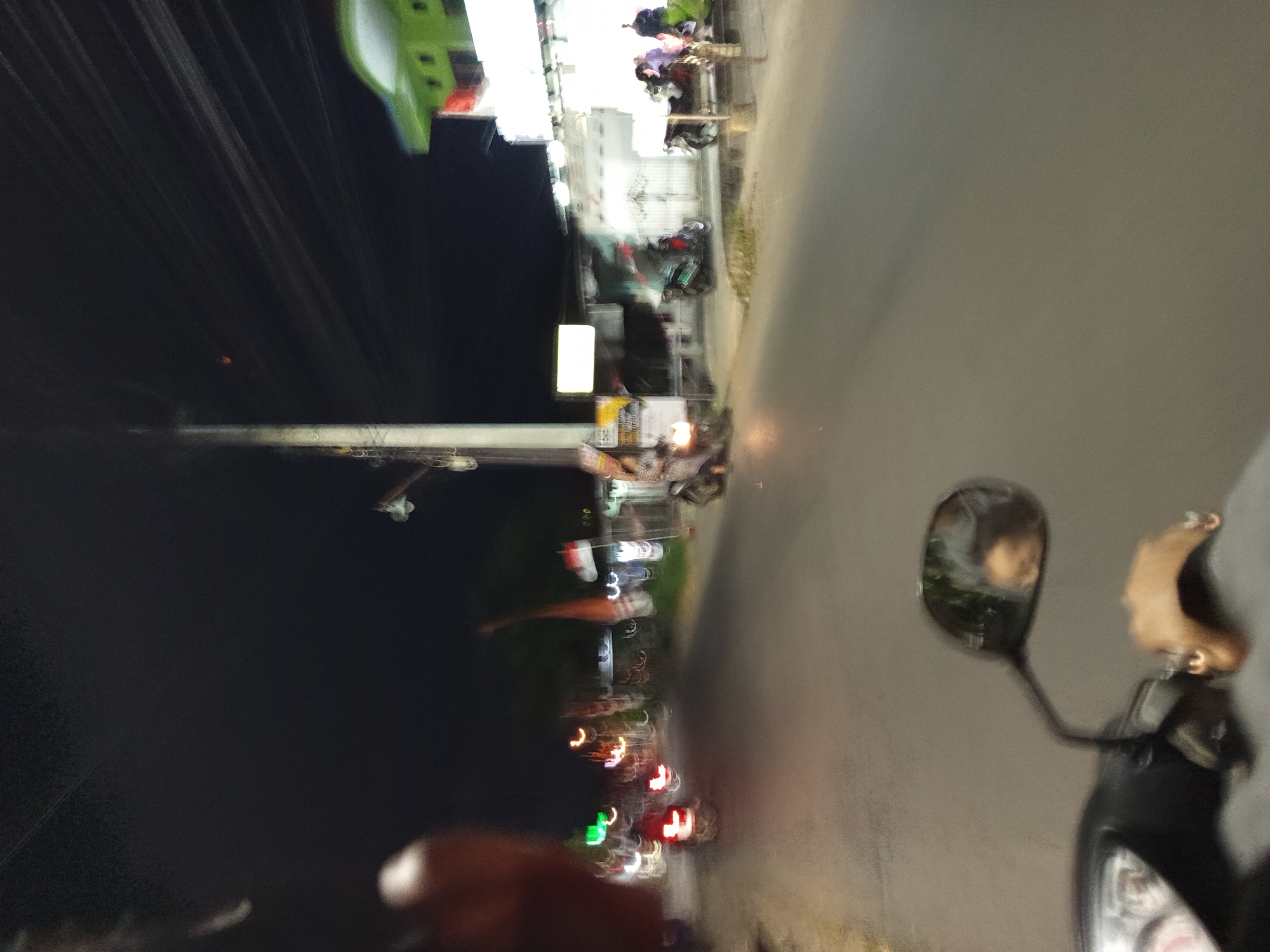 A blurry, nighttime photograph captures a street scene in Punggul, Indonesia. The perspective is from a moving motorcycle, with the handlebars and a rearview mirror in the foreground. The rearview mirror reflects a person's face. The street is illuminated by artificial lights from buildings and street lamps, casting a warm glow on the asphalt. Several motorcycles are visible, some parked and others in motion, with their headlights and taillights streaking due to the camera's motion. On the right side of the street, a building with a green roof and white walls is partially visible. Indonesian flags are displayed in front of some establishments, indicating a national or local celebration. The overall impression is that of a busy street at night, with various activities taking place.