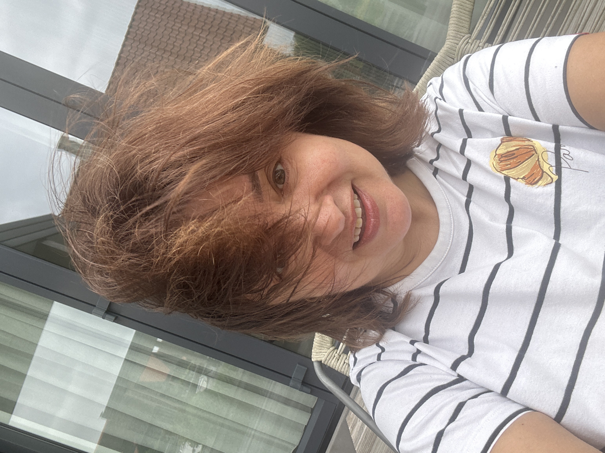 In Ossenzijl, Netherlands, a woman with reddish-brown hair is photographed from the chest up, seated outdoors in what appears to be a modern home with large windows and a tiled roof visible in the background. Her hair is windswept, obscuring part of her face, but her right eye and mouth are visible. She is smiling, with her teeth showing. She is wearing a white t-shirt with black horizontal stripes and a graphic of a croissant and the word "Paris" printed on the left side of her chest. The setting suggests a casual, possibly residential environment, likely on a patio or balcony, given the chair and wooden decking. The overall lighting is natural and diffused, indicating an overcast day.