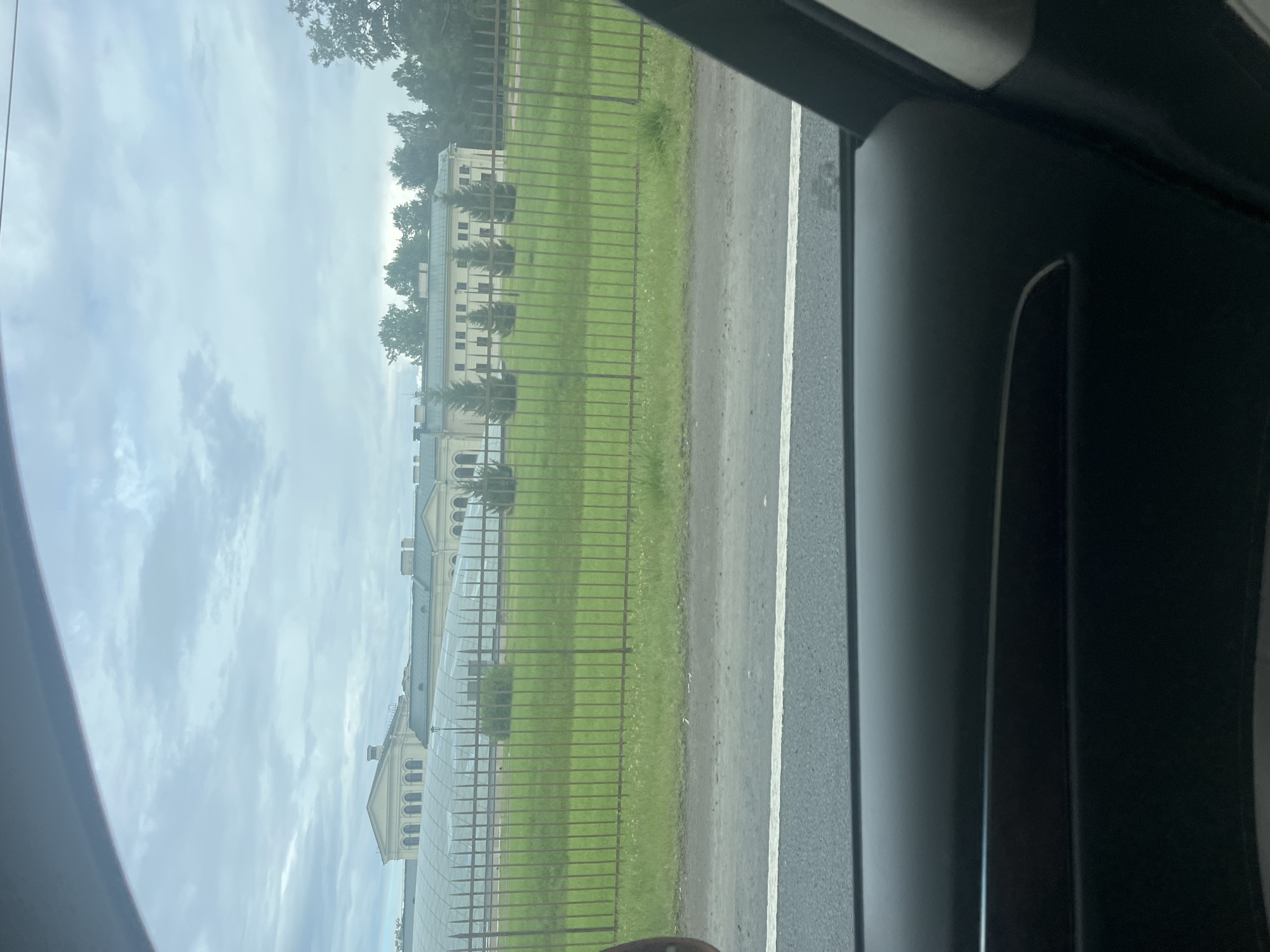 The image captures a view from inside a vehicle looking out at a long, light-colored building with a series of arched windows and a mansard roof. In front of the building is a metal fence with pointed finials, behind which is a grassy area with several conical evergreen trees. Beyond the fence and trees, a road with a white line is visible, and the foreground shows the interior of a car's black dashboard and door panel. The sky is overcast with clouds. The scene is located in Peterhof, Russia, likely near a park or public institution.