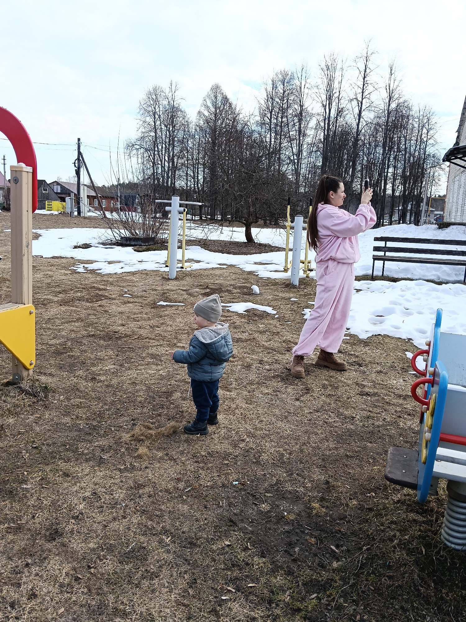 Outside on a playground in Krasny Mayak, Russia, a young child and a woman are present. The child, dressed in a blue jacket, dark pants, and a light grey knit hat, stands near a patch of dead grass in a playground with red and yellow equipment. To his right, a woman in a pink tracksuit and brown boots is taking a picture with her phone. The ground is covered in patchy brown grass with remnants of snow, and leafless trees are visible in the background. In the background, there are residential houses and a wooden bench near the remains of the snow cover.
