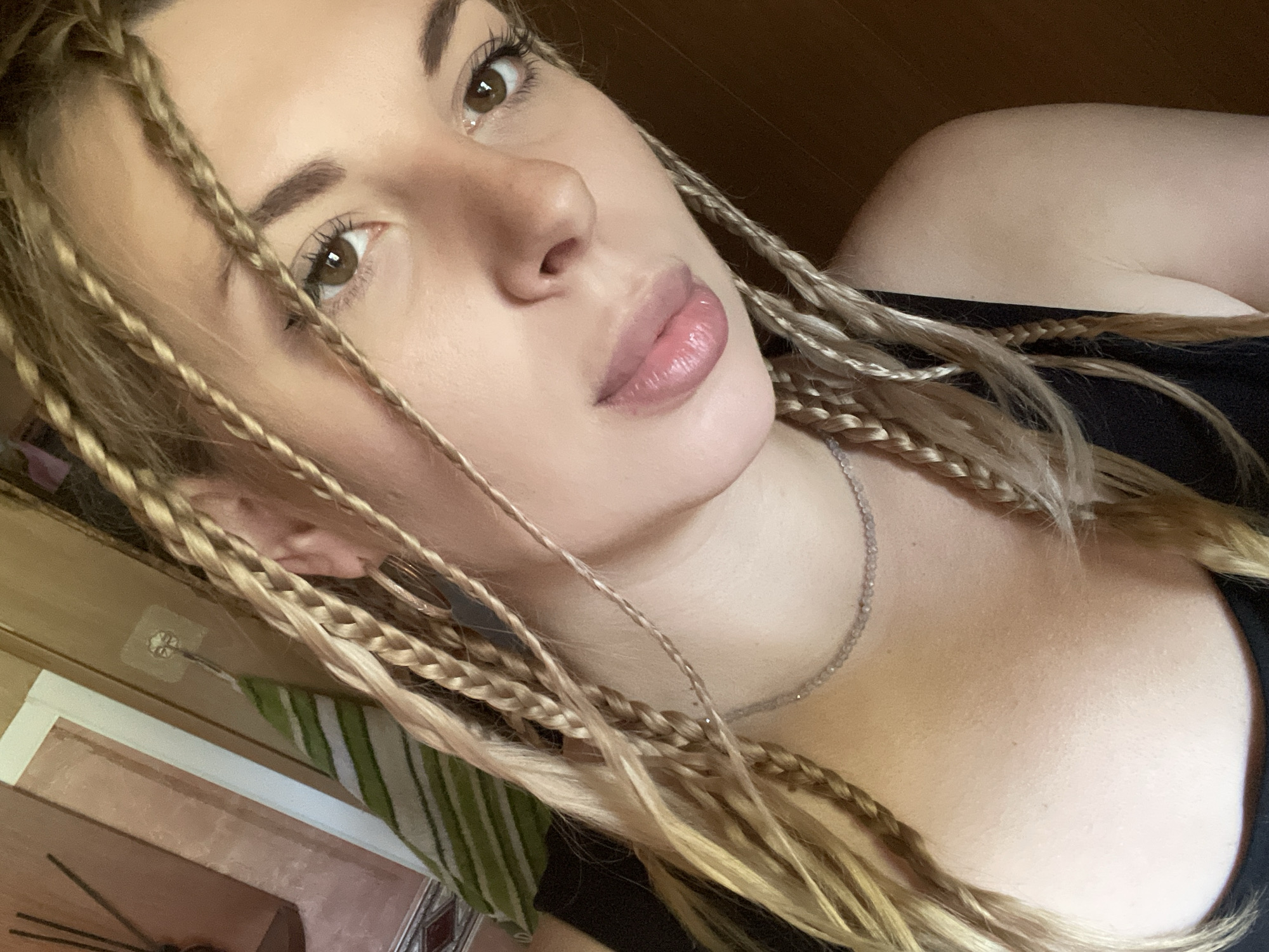 This is a close-up selfie of a young woman with blonde braided hair, taken indoors. She has brown eyes and is wearing a black top and a delicate silver necklace. The background appears to be a wall with some shelving and a green striped object. The lighting suggests it is daytime, possibly near a window. There is no indication of the specific location in Shcherbinka, Russia beyond the context provided.