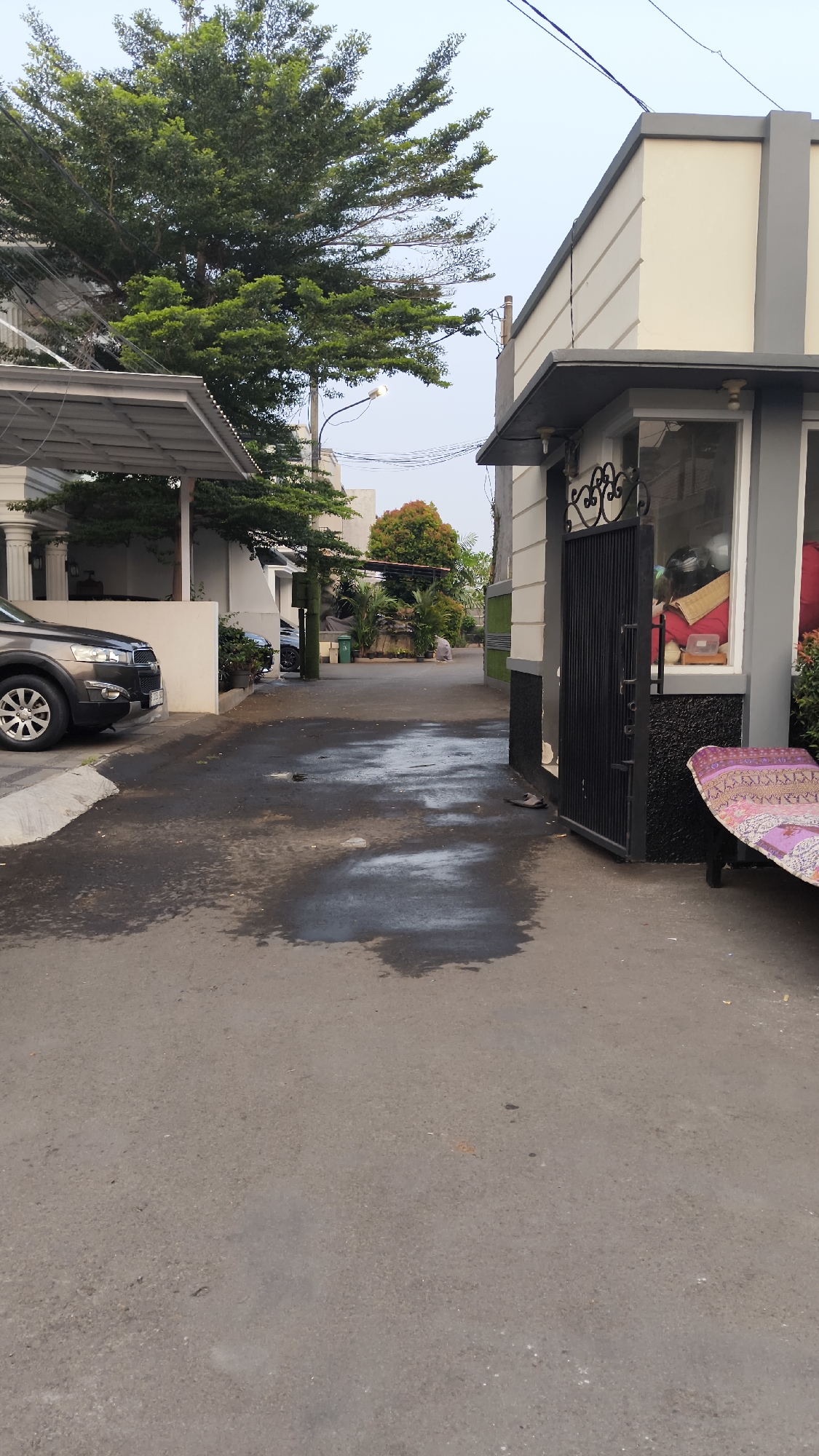The image shows a street in South Jakarta, Indonesia. On the left, a dark gray SUV is parked under a carport with a white roof and pillars. Behind the SUV, a black car is visible. The street itself is paved and appears wet in places, with puddles reflecting the overcast sky.
On the right side of the street, a house with a light beige exterior and dark gray trim is prominent. A black wrought-iron gate is open, leading into what appears to be a small entryway or covered area. Inside the glass-fronted area of the house, items like a red cushion, a helmet, and other indistinct objects are visible. In front of this house, a low bench or seating area is covered with a patterned textile.
Further down the street, a person in light-colored clothing is walking away from the camera. Several trees are scattered throughout the scene, including a large, leafy tree on the left and a tree with red foliage in the distance. The sky is a pale blue, suggesting it is either early morning or late afternoon. Utility poles and wires are visible against the sky. The overall environment suggests a residential street in a tropical climate.