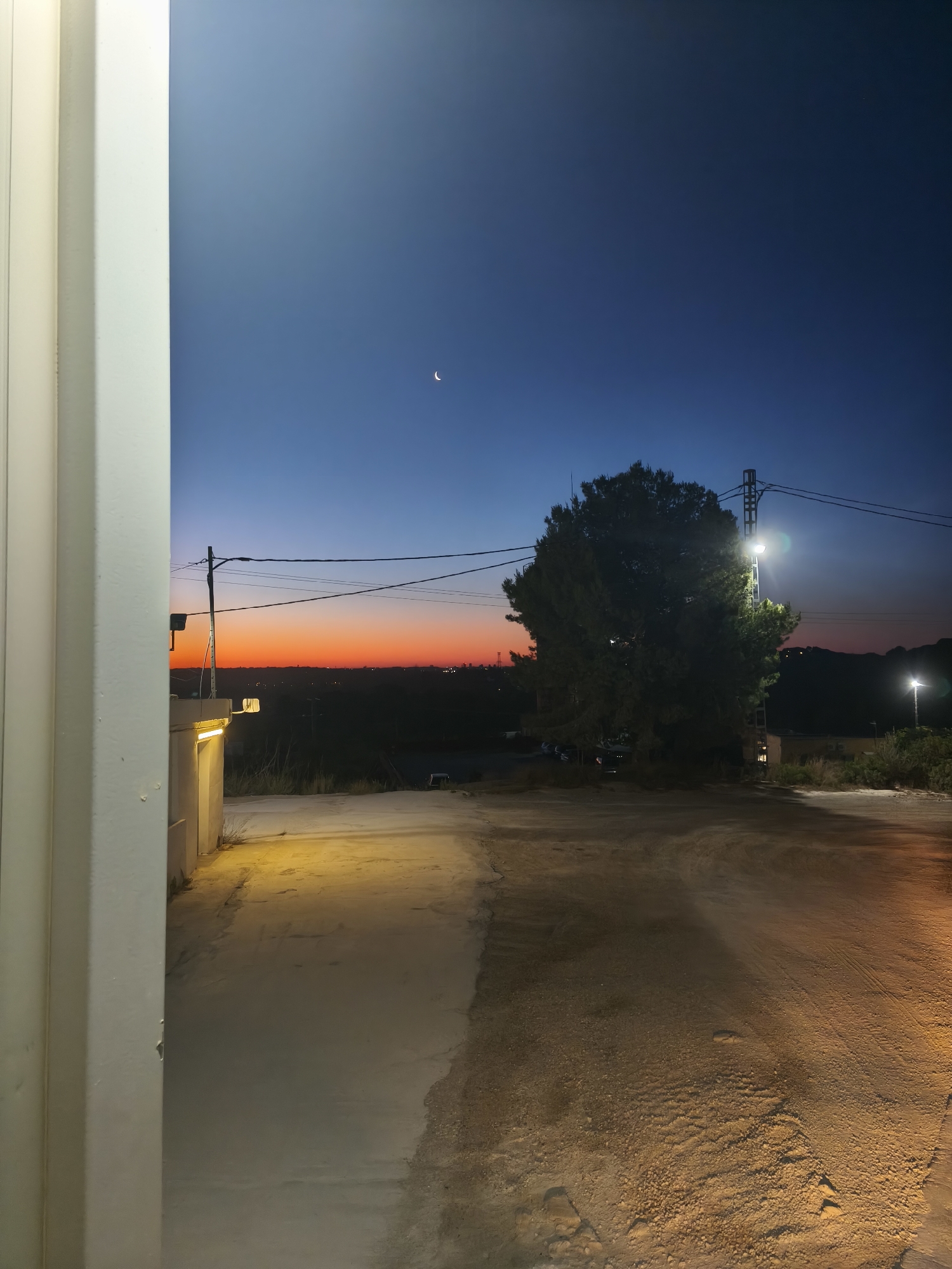 The image depicts an outdoor scene at dusk or twilight. A crescent moon is visible in the upper center of the frame against a dark blue sky that transitions to orange at the horizon. A large tree is centrally located in the middle ground, illuminated by a nearby streetlight on a metal pole. Power lines run across the sky. The foreground shows a dirt or gravel road illuminated by lights, one visible on the right. A partially visible white wall or structure is on the left side of the frame. The location is likely Riba-roja de Túria, Spain, as specified. No people are visible. The environment appears rural or suburban.