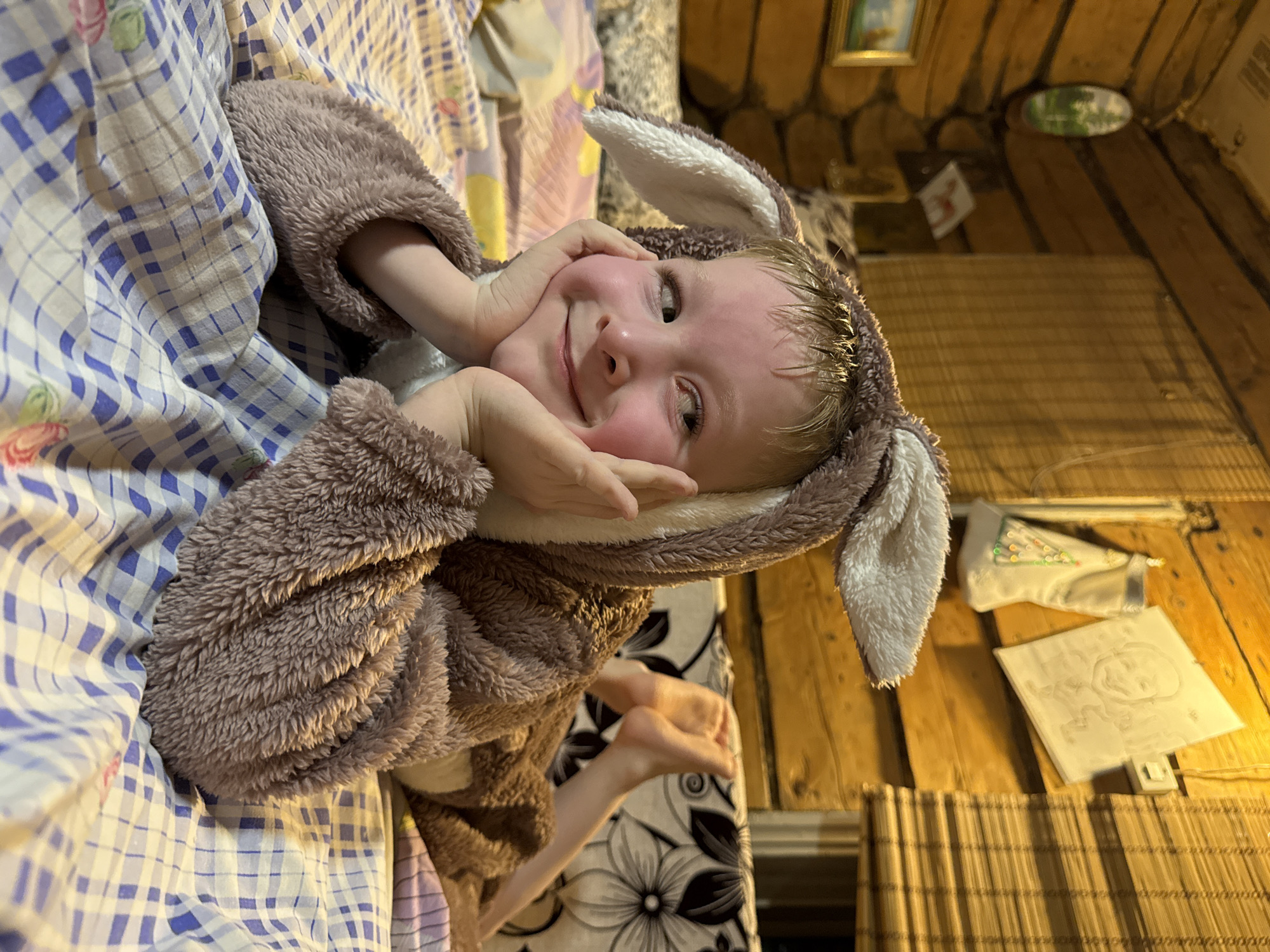 A young child, likely a toddler, is prominently featured, lying down and smiling widely directly at the camera. The child is dressed in a cozy, full-body animal costume made of fluffy brown and white material, complete with visible ears on the hood, suggesting a bunny or bear. Their hands are placed playfully under their chin, highlighting their rosy cheeks and cheerful expression.
The child is positioned on a bed covered with a blue and white checkered blanket or sheet, with another patterned fabric visible underneath. The background reveals a rustic interior with walls and potentially a ceiling constructed from warm-toned wooden planks or logs. A small framed landscape painting and an oval decorative item adorn the wooden wall.
On a surface to the right, a white, mask-like object with green and red patterns is visible, alongside a square white panel featuring a faint, hand-drawn face and scribbles, with a small switch at its base. A bamboo or rattan mat is partially visible in the lower right. The scene depicts a joyful and playful moment within a cozy, rustic home setting in Nizhnyaya Chermoda, Russia. No discernible text is present in the image.