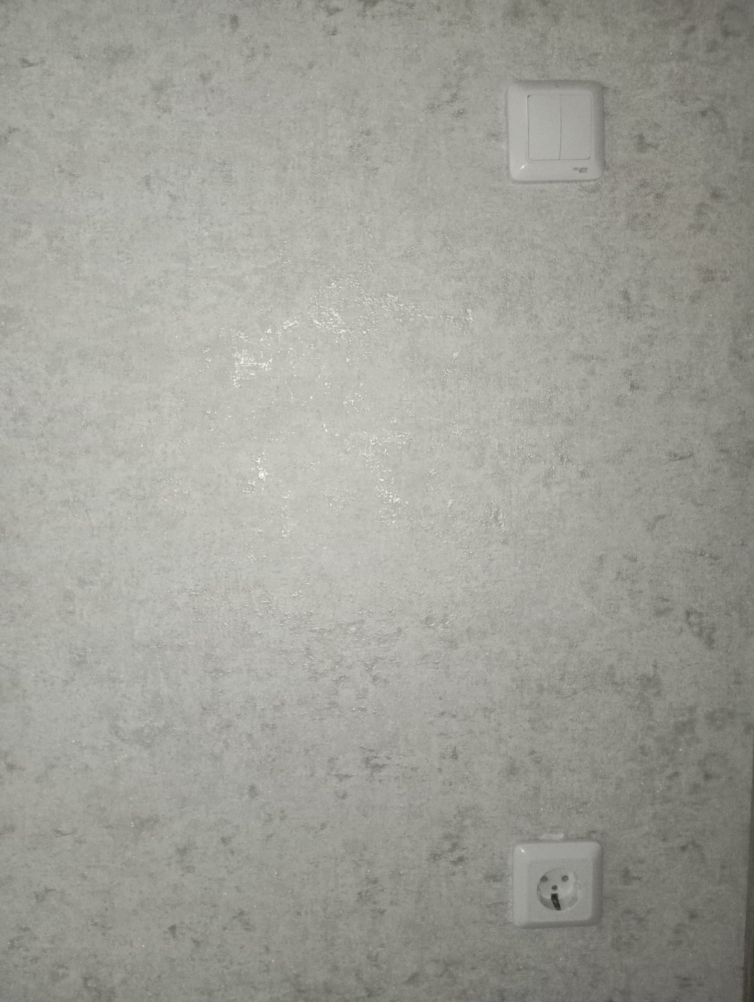 The image shows a light gray, textured wall with two electrical fixtures. In the upper right quadrant, there is a white double light switch. In the lower right quadrant, there is a white electrical outlet with a European-style socket. The wall appears to be wallpapered and has a subtle shimmering effect. The scene is indoors and appears to be a residential or commercial setting. There are no people or other objects visible in the image.