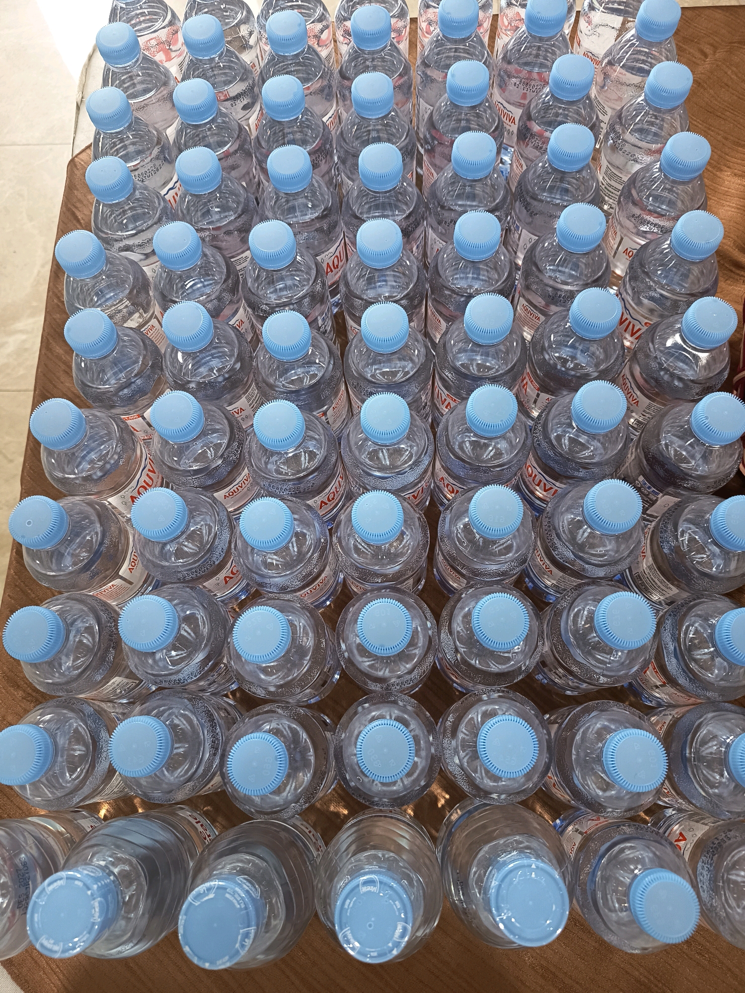 The image shows a top-down view of numerous plastic bottles of water arranged in rows on a brown surface, possibly a table or a mat. The bottles have light blue caps and clear water. Some of the bottles have visible labels with text. The scene appears to be indoors, with the lighting suggesting an artificial light source. There are no people or any other objects visible in the image. The arrangement of the bottles is orderly, with rows and columns. The surface they are placed on has a textured appearance. The image is a static shot of a large quantity of bottled water.