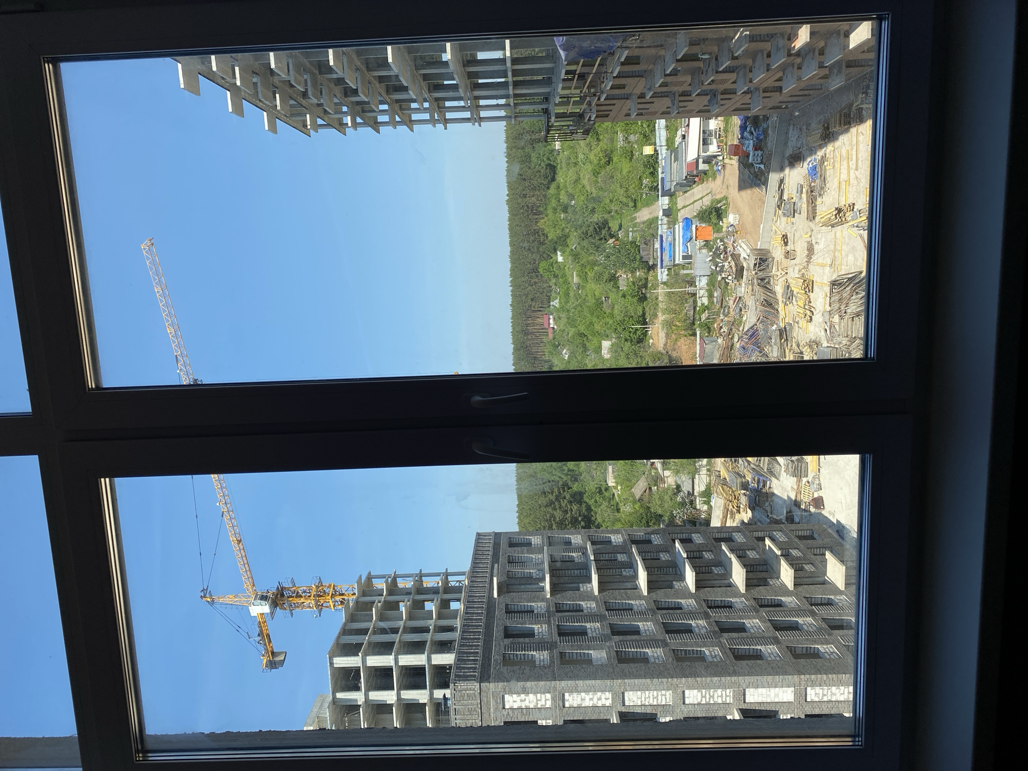 The image, taken from inside a room through a closed window, depicts an ongoing construction site in what is likely a residential area of Izhevsk, Russia. The lower pane of the window frame reveals a high-rise building under construction. A yellow tower crane is visible, actively involved in the building's construction. The building's facade is a mix of exposed concrete and brickwork, showing the early stages of development. The upper pane of the window reveals a different construction site with a partially built structure, a clear blue sky, and a distant treeline suggesting a forest or park area on the outskirts of the city. Various materials, equipment, and construction debris are visible at the base of the unfinished structures, indicating continued construction activity. The scene suggests the ongoing urban development and expansion in Izhevsk, juxtaposing new construction with the natural environment.