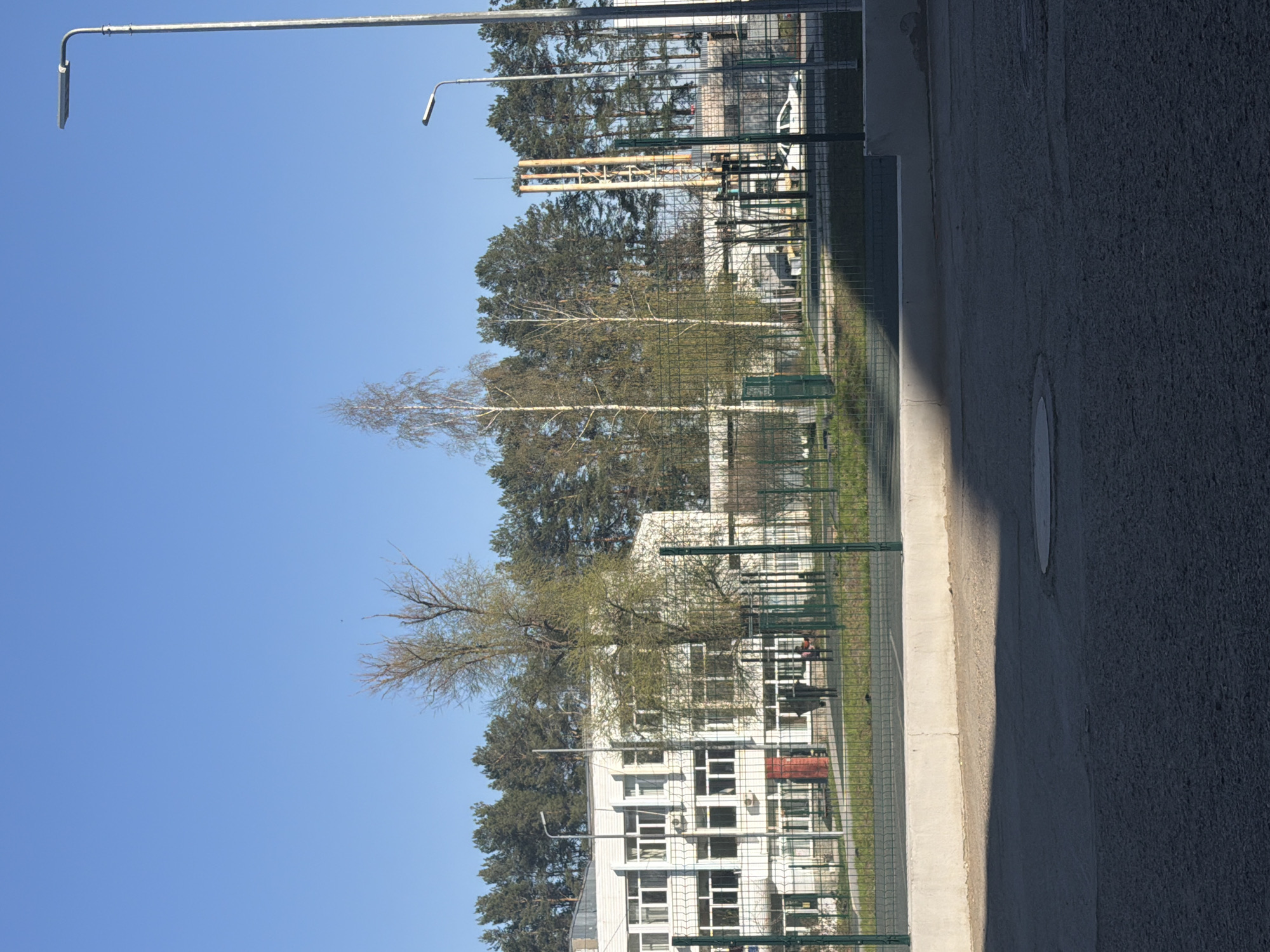 The image shows a street scene in Chaikovsky, Russia. A multi-story white building with numerous windows is partially visible behind a green chain-link fence. Tall trees and utility poles are present. Two people are sitting on a bench in front of the building. The sky is clear and blue. The foreground consists of a concrete sidewalk and asphalt road.