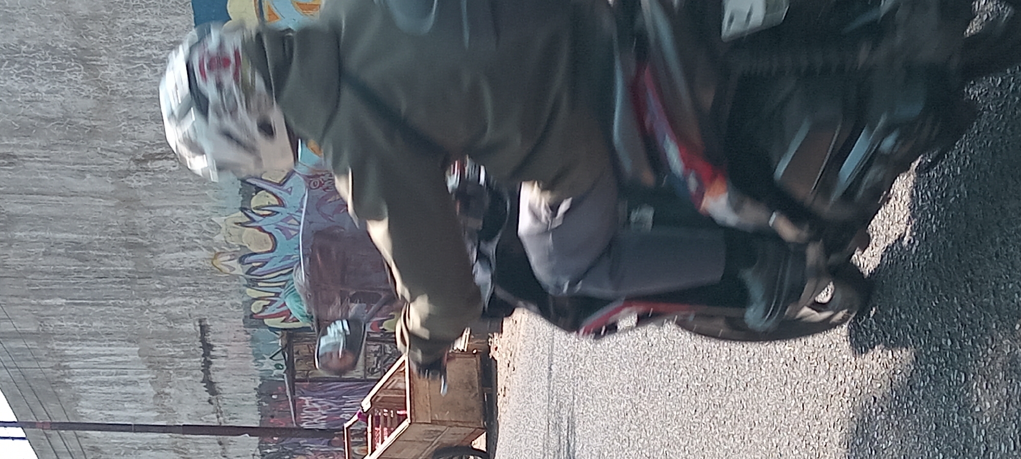 The image depicts a person riding a motorcycle on a paved road, with a concrete wall covered in graffiti as the background. The rider is wearing a green jacket and a helmet, and is holding onto the handlebars. To the left of the motorcycle is a wooden cart with red items inside. The scene appears to be in an urban environment, possibly a street in Tasikmalaya, Indonesia, given the presence of graffiti and the typical urban landscape. The lighting suggests it is daytime, with sunlight casting shadows on the road.
