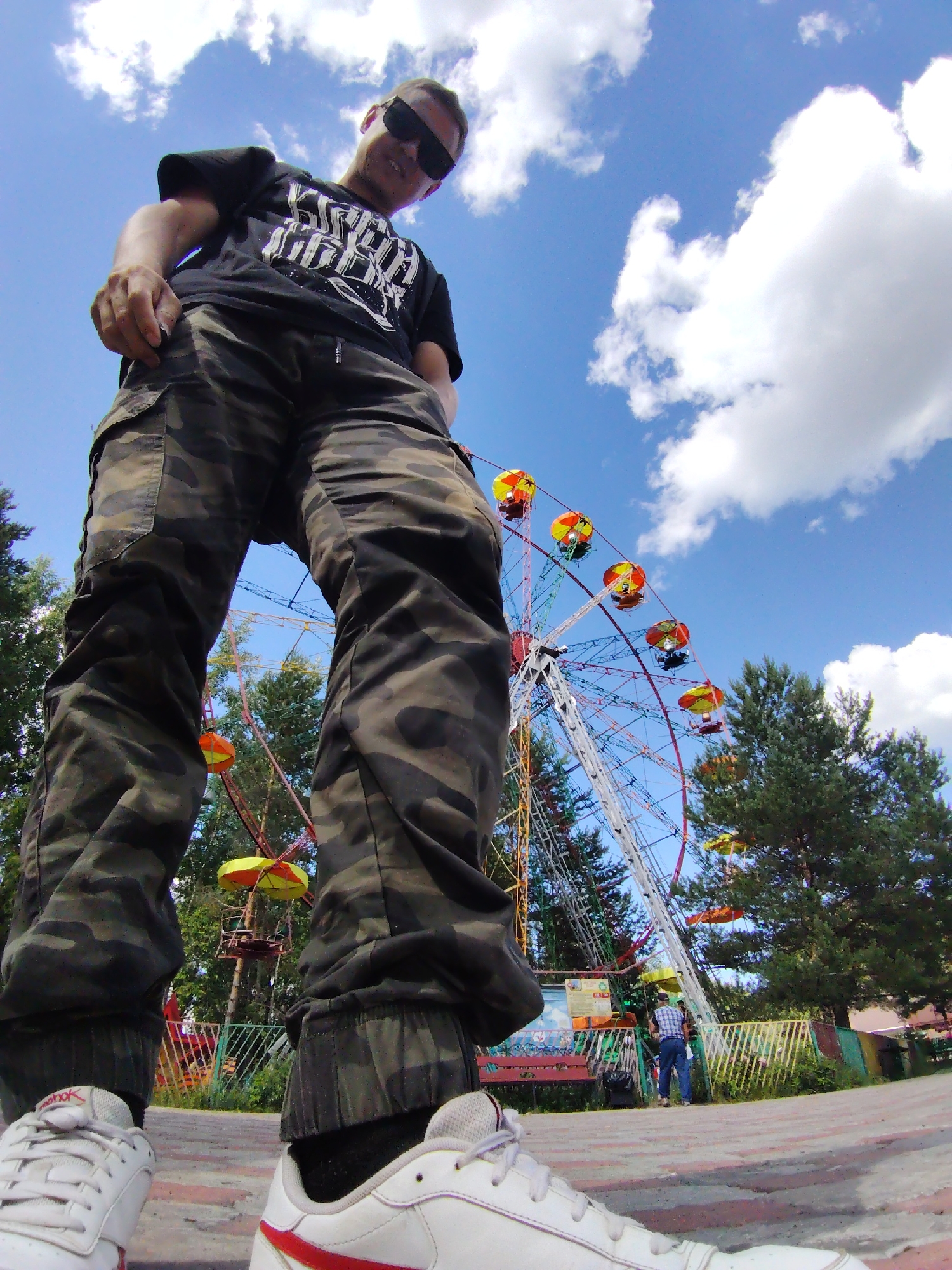 A low-angle, wide-shot captures a person in the foreground, their legs and torso dominating the frame. They wear camouflage pants and white sneakers. Behind them, a Ferris wheel is visible, with colorful gondolas. Trees and a bright blue sky with fluffy white clouds complete the scene.
The image appears to be taken on a sunny day. The presence of the Ferris wheel suggests an amusement park or fairground setting. The person's posture and the camera's low angle create a dynamic and perhaps playful perspective.
In the background, another person is standing near the base of the Ferris wheel, wearing a blue and white checkered shirt and jeans. Several signs are visible on a structure near the Ferris wheel, but the text is not legible. The overall mood is bright and cheerful, typical of a day at an amusement park.
The location is identified as Chaikovsky, Russia. The time of day is likely daytime, given the bright sunlight and clear sky.