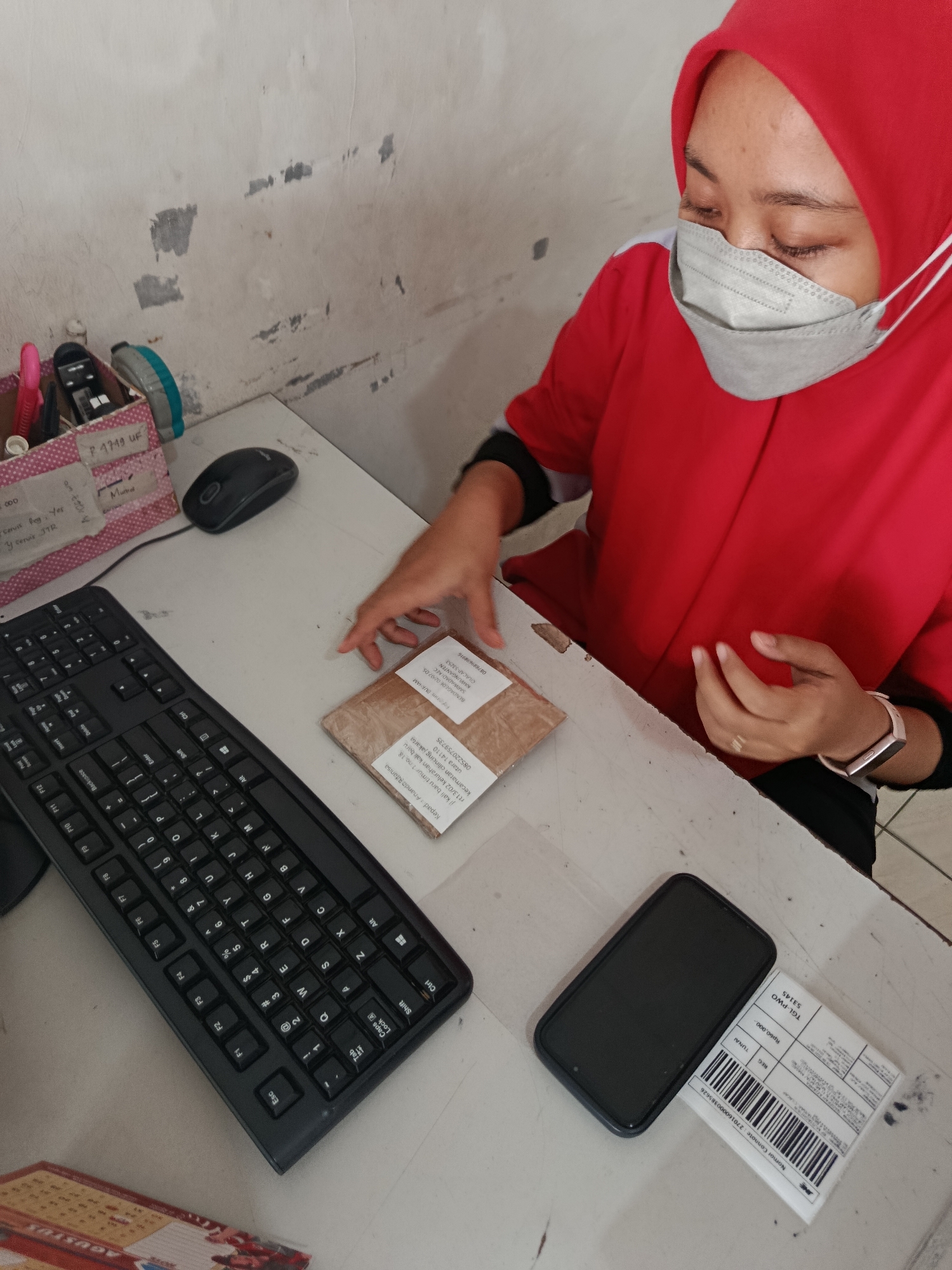A person wearing a red headscarf and a grey face mask sits at a desk, with a keyboard and a mouse in front of them. They are handling a small package wrapped in brown paper. To the right of the package is a black smartphone and a white shipping label with barcodes. A pink and white organizer filled with pens and other items is on the left side of the desk, next to a mouse. The wall behind the desk is textured and appears aged, with patches of peeling paint. The scene suggests a home office or a small business environment in Bantarsari, Indonesia.