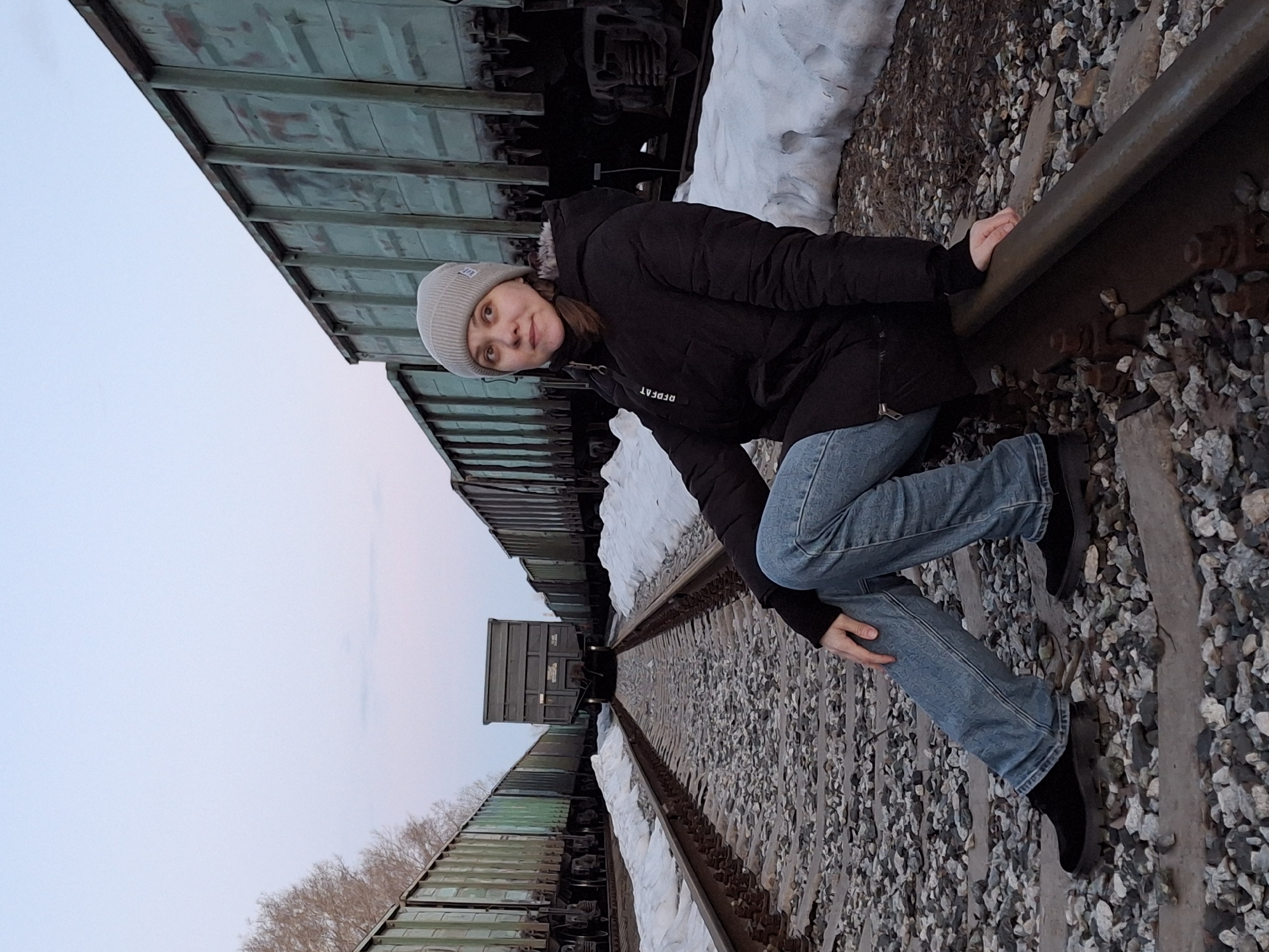 A woman sits on a railroad track. She is wearing a gray knit cap, a black jacket with "REPEAT" printed on it, blue jeans, and black shoes. She is positioned between two tracks, with one hand on each rail. Above her are a series of green freight cars. The ground around the tracks is covered in small stones and patches of snow. The sky is visible in the upper left, a light gray color. The photo was taken in the city of Chaikovsky, Russia.