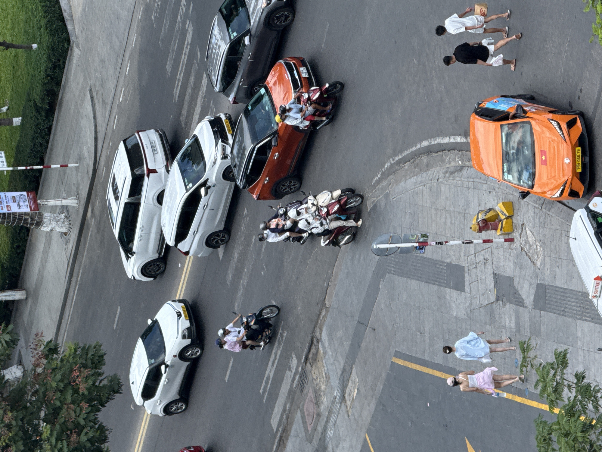 The overhead shot depicts a street scene in Nha Trang, Vietnam, on a paved road with yellow and white lane markings. Several vehicles, including white SUVs, a gray SUV, a red SUV, and a distinctive orange taxi, are on the road. Multiple motorcycles are also present, with riders often wearing helmets and carrying passengers. Two pedestrians, appearing to be women, walk along the sidewalk, one in a light blue top and white shorts, the other in a white top and a pink skirt. A red fire hydrant and yellow barriers are visible on the sidewalk. The environment includes trees and greenery, suggesting an urban setting.