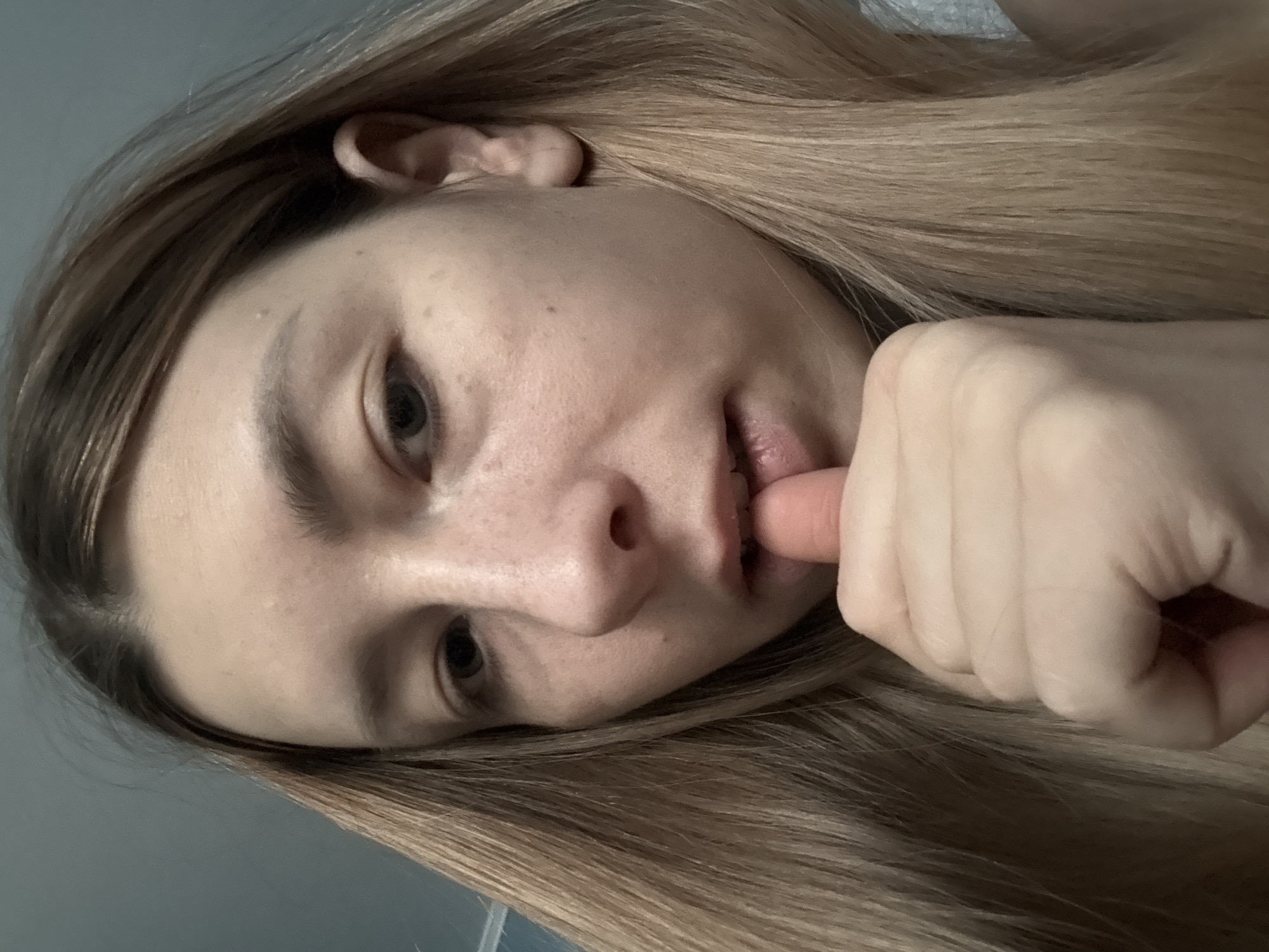 A close-up, angled portrait shows a person with fair skin and light brown or blonde hair, partially obscuring the face. The person's left index finger is placed near their mouth, touching the lower lip. The background appears to be a neutral, possibly grayish, wall. No details suggest a location in Moscow, Russia.