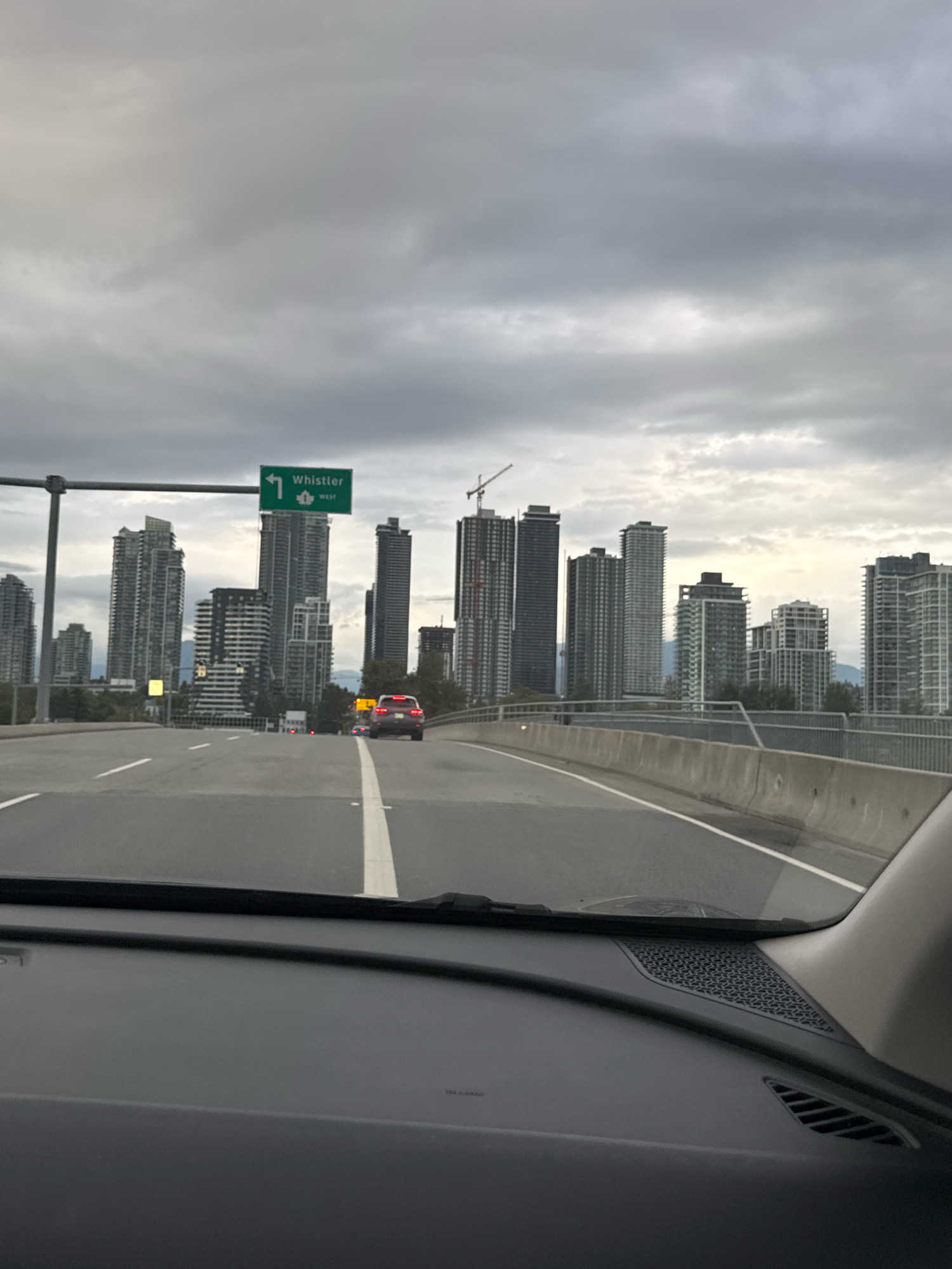 The image shows a view from inside a vehicle looking out at a highway and a city skyline. A green highway sign with white text indicating "Whistler" and an arrow pointing left is visible in the upper left quadrant of the image. The city skyline consists of numerous tall, modern buildings, some of which appear to be under construction. The sky is overcast with gray clouds. The highway has multiple lanes, and a red SUV is driving in one of the lanes. The interior of the vehicle is visible in the foreground, including the dashboard. The scene is likely captured in Burnaby, Canada, as indicated by the Whistler highway sign, which is a common direction for drivers heading north from the Vancouver area.