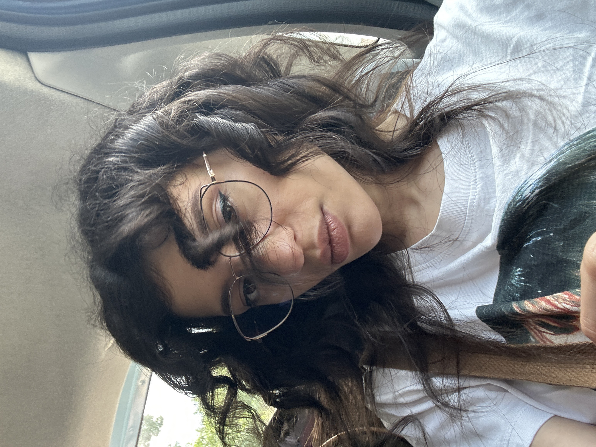 The image is a close-up, low-angle, outdoor, slightly over-the-shoulder selfie of a young woman with long, dark, wavy hair. She is wearing a white t-shirt and delicate, round-framed glasses. Her head is tilted, and she is looking directly at the camera with a relaxed, slightly amused expression. Her lips are slightly parted, revealing a hint of a smile. The lighting suggests it is daytime, likely overcast or in a shaded area, as there are no harsh shadows.
The background is a blurred interior of a vehicle, with the car's interior structure visible in the upper left corner. Through the car window on the right, glimpses of greenery can be seen, suggesting a location with trees. The overall impression is casual and spontaneous, possibly taken while traveling in a car. The woman's hair is partially obscuring her face, adding a sense of movement and naturalness. There is no visible text in the image. The context provided is Sofia, Bulgaria.
