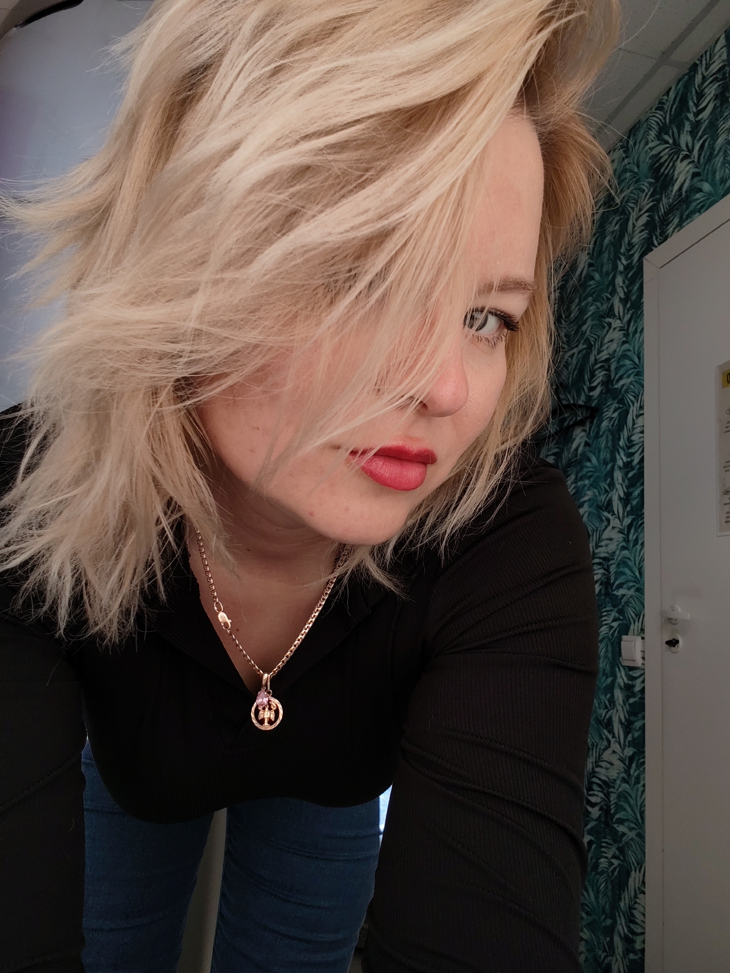 The image is a selfie of a young woman. She has blonde, layered hair that falls across her face, partially obscuring her left eye. She has fair skin and is wearing red lipstick. She is wearing a black, long-sleeved ribbed shirt and a gold necklace with a round pendant. She appears to be wearing blue jeans. The background features a wall covered in a green and white tropical leaf pattern. A white door with a door handle and a framed sign are visible in the background. It is not possible to determine the location of the photo or its coordinates in Izhevsk, Russia based on the provided information.
