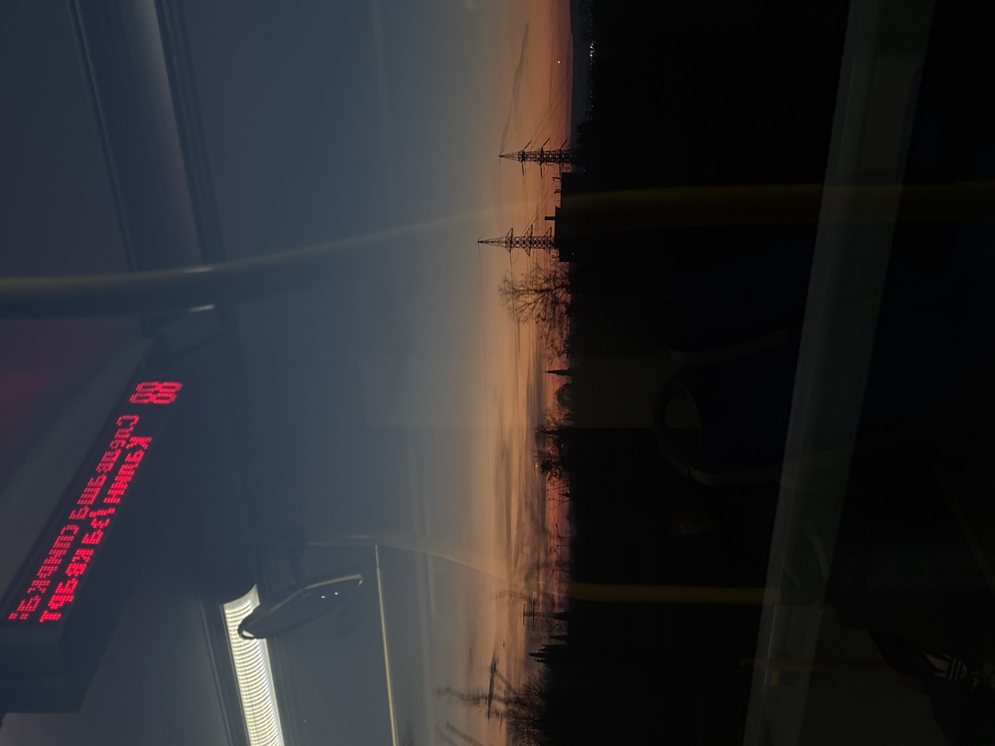 The image appears to be taken from inside a bus, likely in Varna, Bulgaria, as evidenced by the Cyrillic script on the electronic display showing the route number "88" and other text. The view through the window captures a twilight or sunset scene. Overhead power lines are visible silhouetted against the fading sky. The landscape appears dark, suggesting a residential or industrial area beyond the road. Reflections are present on the window, blurring the view of the outside with the interior of the bus. Part of a metal handrail is seen inside the bus, confirming the location. The overall impression is that of a bus ride at dusk or dawn, with the exterior landscape visible through the window, partially obstructed by the interior of the bus and its reflections.