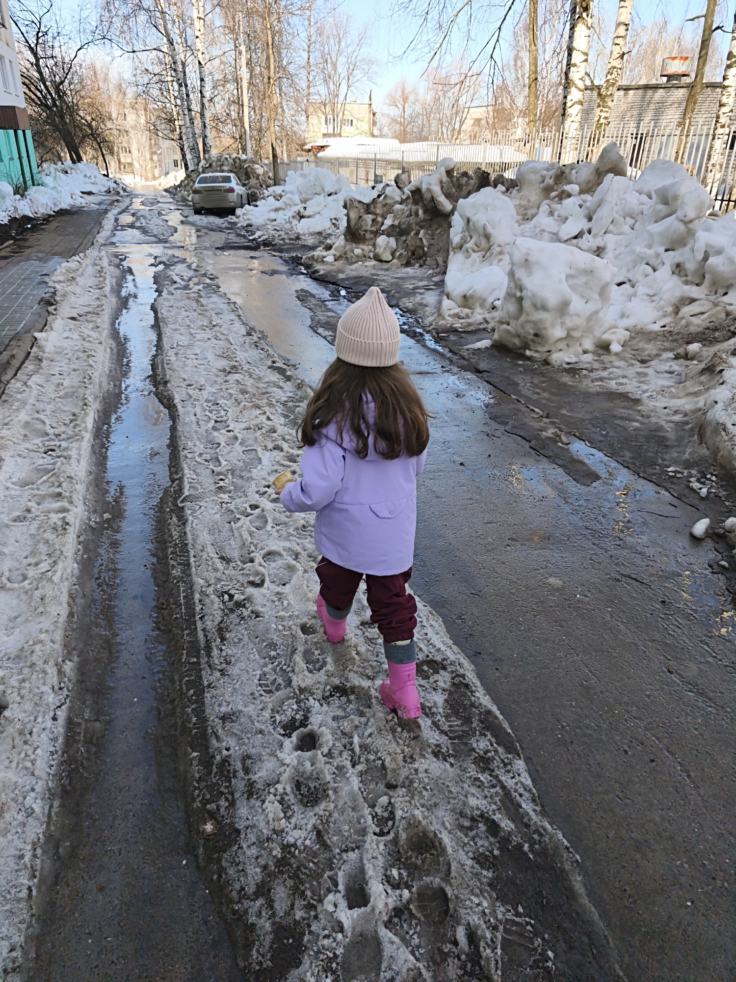 The image depicts a partially cleared residential street in Chaikovsky, Russia, under thawing conditions. A young child, viewed from behind, walks on a slushy, snow-covered portion of the asphalt road. The child is dressed in a light pink beanie, a light purple jacket, maroon pants, and bright pink boots, holding a light-colored object in their right hand. The street itself is degraded, exhibiting melting snow, standing water puddles, and visible tire tracks. Large, dirty piles of snow line both sides of the road. On the left, a multi-story building with a teal lower facade is visible. In the mid-ground, a light-colored sedan is parked on the left shoulder, partially obscured by snow and bare deciduous trees, including birch. Further along the right side of the street, beyond a fence and snow piles, a low building with a dark roof and a red cylindrical object on top is present. The sky is clear and blue, and the trees are bare, indicative of late winter or early spring.