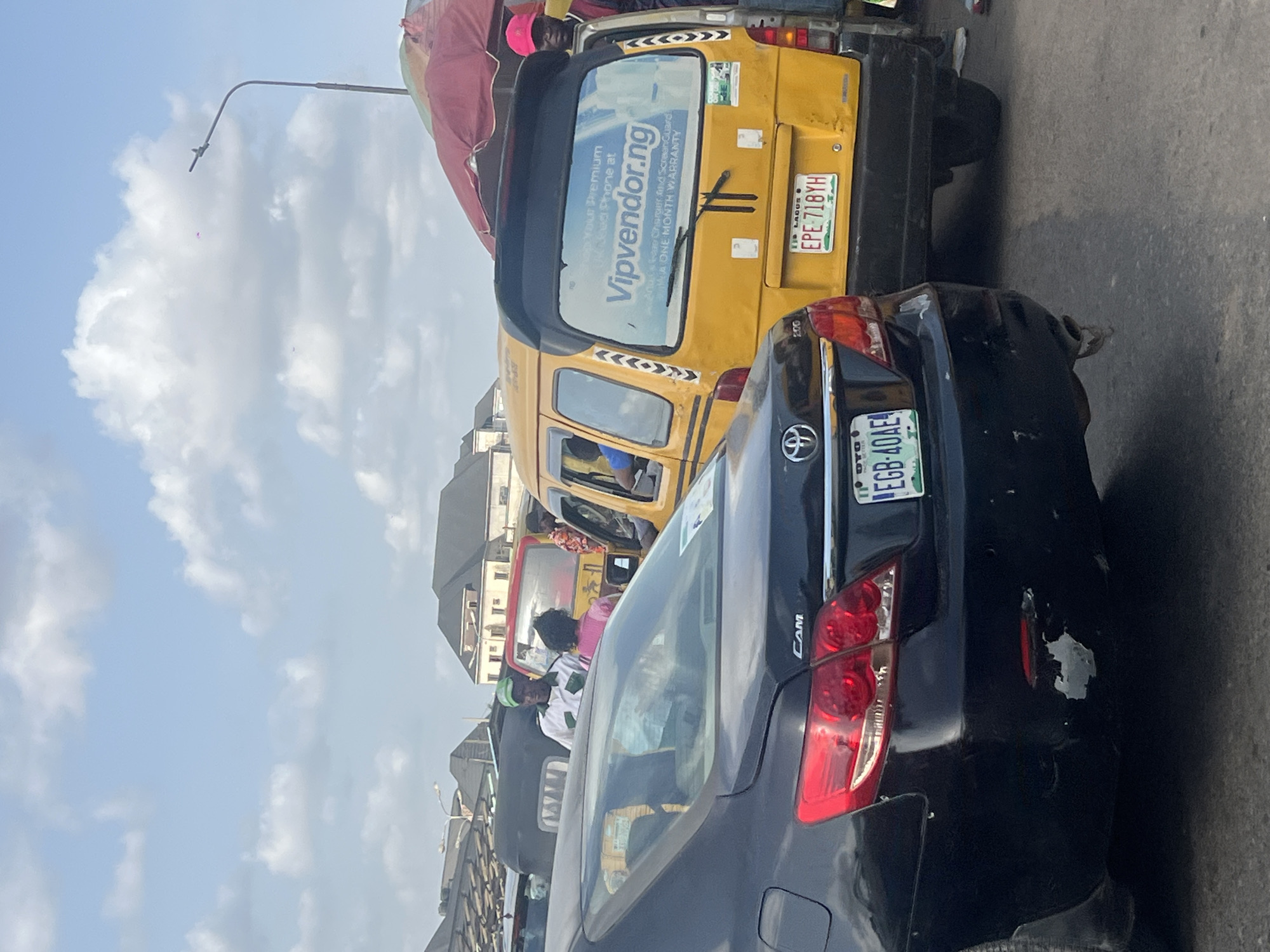 The image captures a congested street in Egan, Nigeria. A blue Toyota Camry with license plate EGB-40AE is prominently positioned, showing damage to its rear. A yellow commercial bus with license plate EPE-718YH is directly behind the Camry. The bus has an advertisement for Vipvendor.ng on its rear window. The presence of numerous other vehicles and people suggests heavy traffic. The background includes buildings and a sky with scattered clouds.