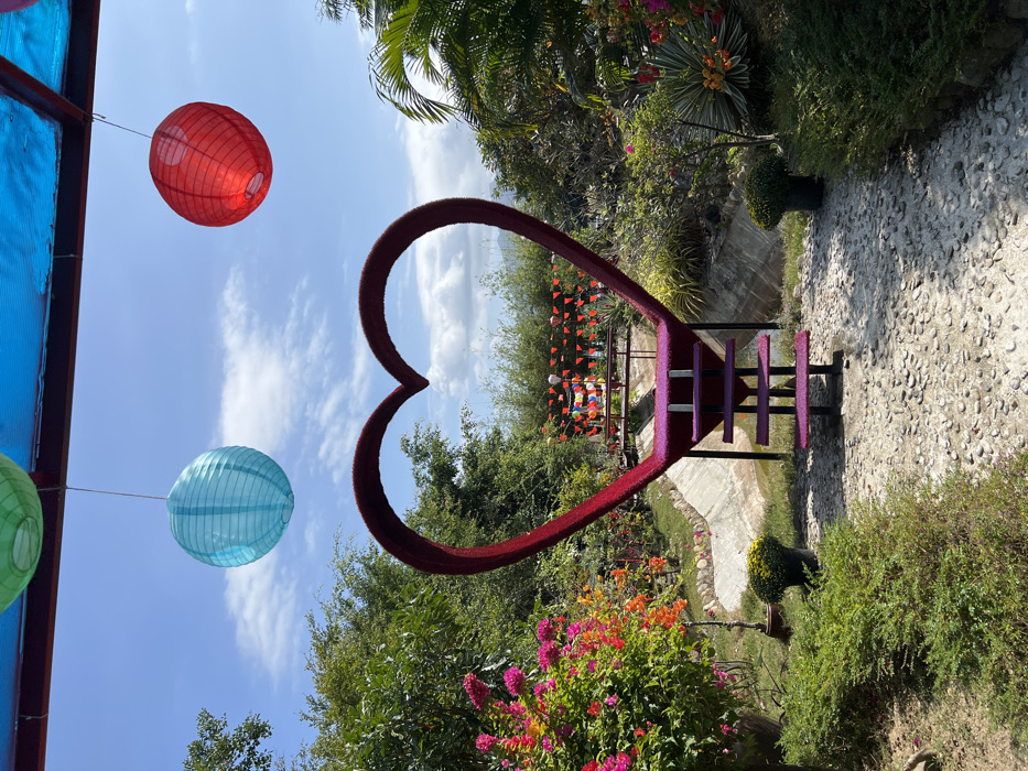 This image captures a vibrant and picturesque outdoor scene, likely a garden or park, under a bright, clear sky. The photo is rotated 90 degrees clockwise from its natural orientation.
The central and most prominent feature is a large, dark red, heart-shaped archway. This archway appears to be covered in a textured material, possibly artificial flowers or a soft fabric, giving it a slightly fuzzy appearance. Inside the heart frame, there is a small platform or seating area with purple steps, which is further decorated with numerous small, colorful triangular pennant flags, suggesting a festive or romantic photo opportunity spot.
Hanging in the upper left of the frame, against the bright blue sky, is a spherical bright red paper lantern. Below it, a light blue paper lantern of the same design is suspended, with a partial view of a green lantern to its left. These lanterns add to the decorative and festive ambiance.
The scene is abundant with lush greenery. Various trees and bushes fill the background and mid-ground. In the lower center and right, there are vibrant clusters of blooming flowers in shades of pink, orange, and yellow, adding rich splashes of color to the landscape. To the right of the heart structure, a light-colored pebbled path stretches into the distance, bordered by a narrow concrete channel or ditch.
The sky is a clear, brilliant blue with scattered, wispy white clouds, indicating fair weather. The entire scene is brightly illuminated by natural sunlight, casting visible shadows, particularly on the pebbled path, suggesting it is daytime. In the distant background, more green foliage and what might be hills or mountains are visible.
There is no visible text in the image.