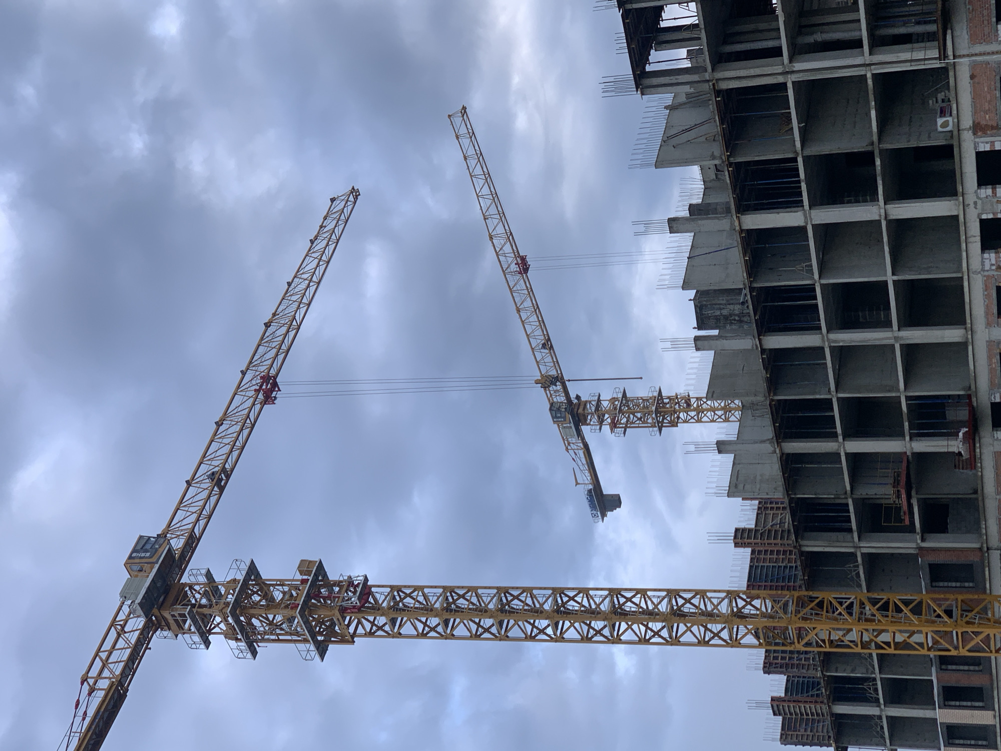 An exterior, slightly upward-angled medium shot shows a construction site in Stavropol, Russia, dominated by two tall yellow tower cranes against a cloudy, overcast sky. The crane on the left is a prominent, full-height structure with a red counterweight section visible at the top. Its long boom extends horizontally across the upper portion of the frame. To the right, a second, partially visible tower crane stands with its boom angled upwards and to the right. In the foreground and midground, the skeletal framework of a multi-story concrete building is under construction. The building features rows of concrete columns and floor slabs, with rebar sticking up from the upper levels, awaiting further construction. Some sections of the building appear to have concrete forms in place. The scene is devoid of visible people. The overall impression is one of active construction in an urban environment under a somber sky.