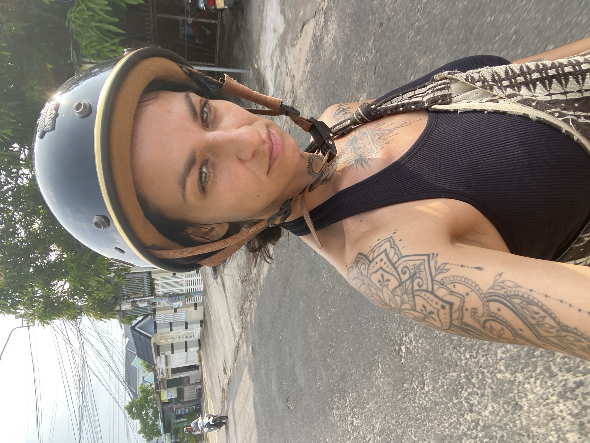 The image shows a woman with a helmet on, on a motorcycle, in Mũi Né, Vietnam. The woman is looking at the camera. She has tattoos on her arms and chest. She is wearing a black tank top and a patterned bag. The background shows a street with houses and trees. There is another motorcycle in the distance. The sky is overcast. The lighting suggests it is daytime.